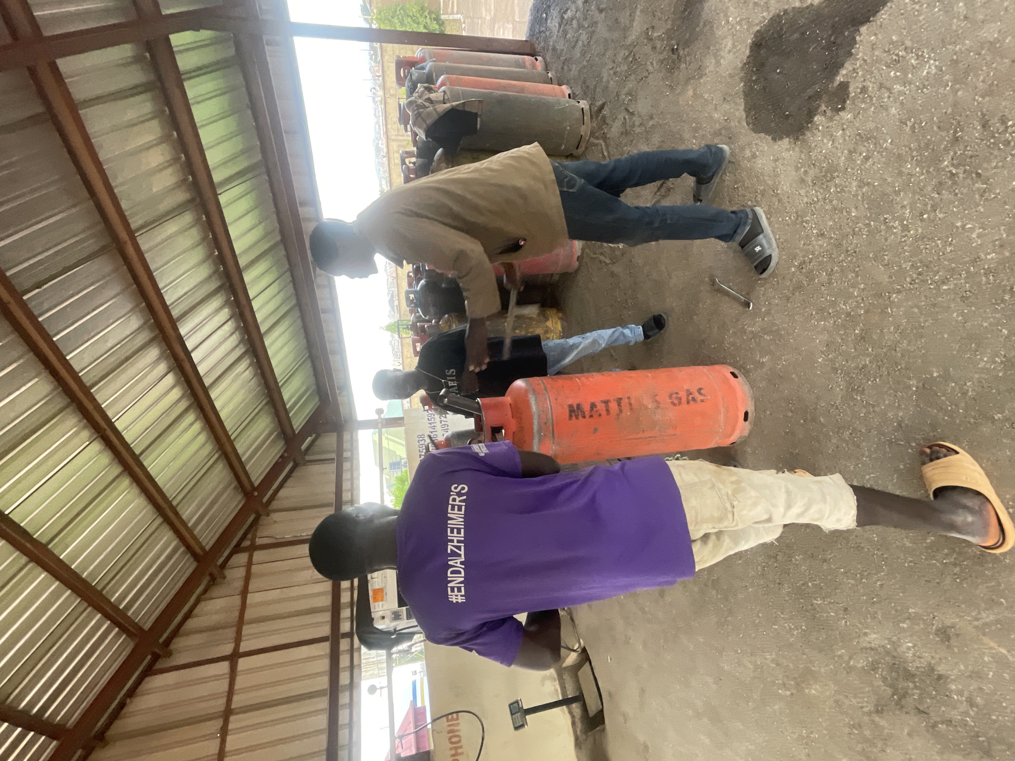 The image depicts a group of individuals in an outdoor, covered area with a corrugated metal roof supported by a metal frame. The ground is a mixture of gravel and dirt. Several large, orange gas cylinders are visible, some stacked in rows and others lying horizontally.
In the foreground, a person wearing a purple t-shirt with the text "#ENDALZHEIMER'S" printed vertically in white on the back is standing. They are also wearing light-colored shorts. Next to them, a red gas cylinder with "MATTILA GAS" printed on it is positioned horizontally. Further back, another person in a black t-shirt and blue jeans is interacting with a gas cylinder, holding a tool that appears to be a wrench. Another individual in a tan jacket and blue jeans is also present, seemingly observing or assisting.
The lighting suggests it is daytime, possibly overcast or in a shaded area due to the roof. The presence of multiple gas cylinders indicates a commercial setting, likely a gas refill or distribution point. The overall atmosphere seems to be one of work or a task being performed, with the individuals focused on their actions. No specific emotions are clearly discernible from their body language. The text on the t-shirt "#ENDALZHEIMER'S" points to a potential cause or awareness campaign being supported. The location context of Jos, Nigeria, suggests this scene is situated in that region.