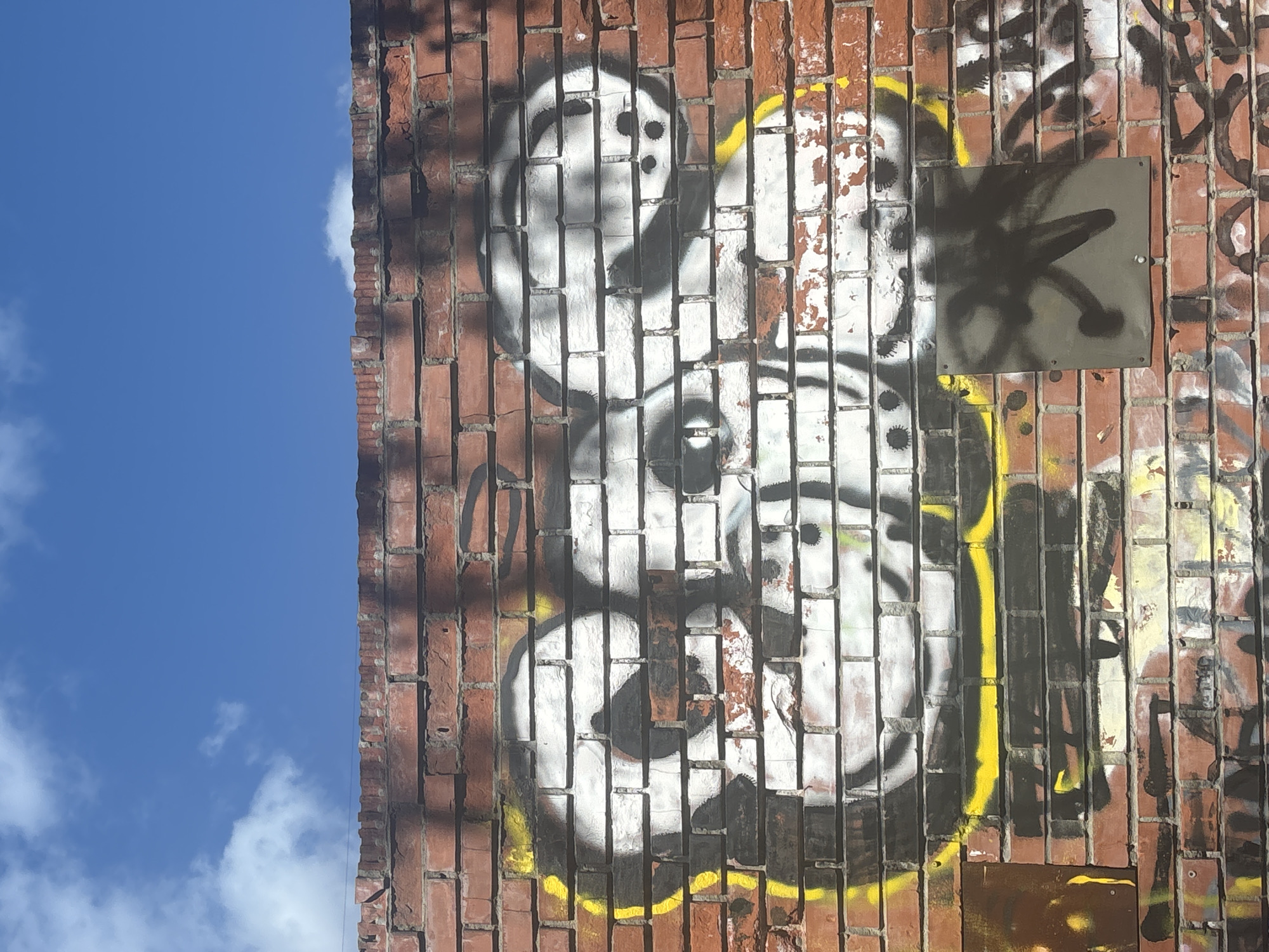 The image shows a brick wall with graffiti. The graffiti is predominantly white with black and yellow accents. The brick wall appears to be aged and shows signs of wear and tear. Above the wall, a bright blue sky with scattered white clouds is visible. The overall impression is of an urban outdoor setting. The location is stated to be Novosibirsk, Russia. The graffiti appears to be a stylized word or symbol, rendered in a bold, graffiti art style. There are no people or animals visible in the image. The lighting suggests it is daytime, with shadows cast on the wall from an unseen source. The wall itself is made of red brick, and some of the bricks are damaged or discolored. The graffiti covers a significant portion of the wall's surface. There is also a dark gray square object attached to the wall with screws, which is partially covered by graffiti.