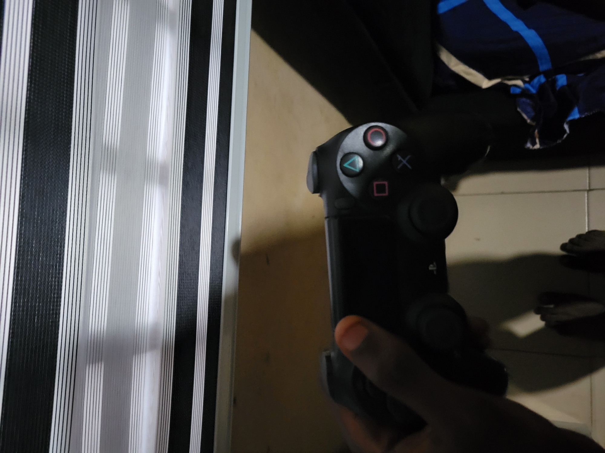 A person is holding a black PlayStation 4 controller in their left hand. The controller has the standard button layout, including the triangle, circle, cross, and square buttons, as well as two analog sticks. The background consists of horizontal blinds and a wooden surface. The lighting suggests an indoor environment.