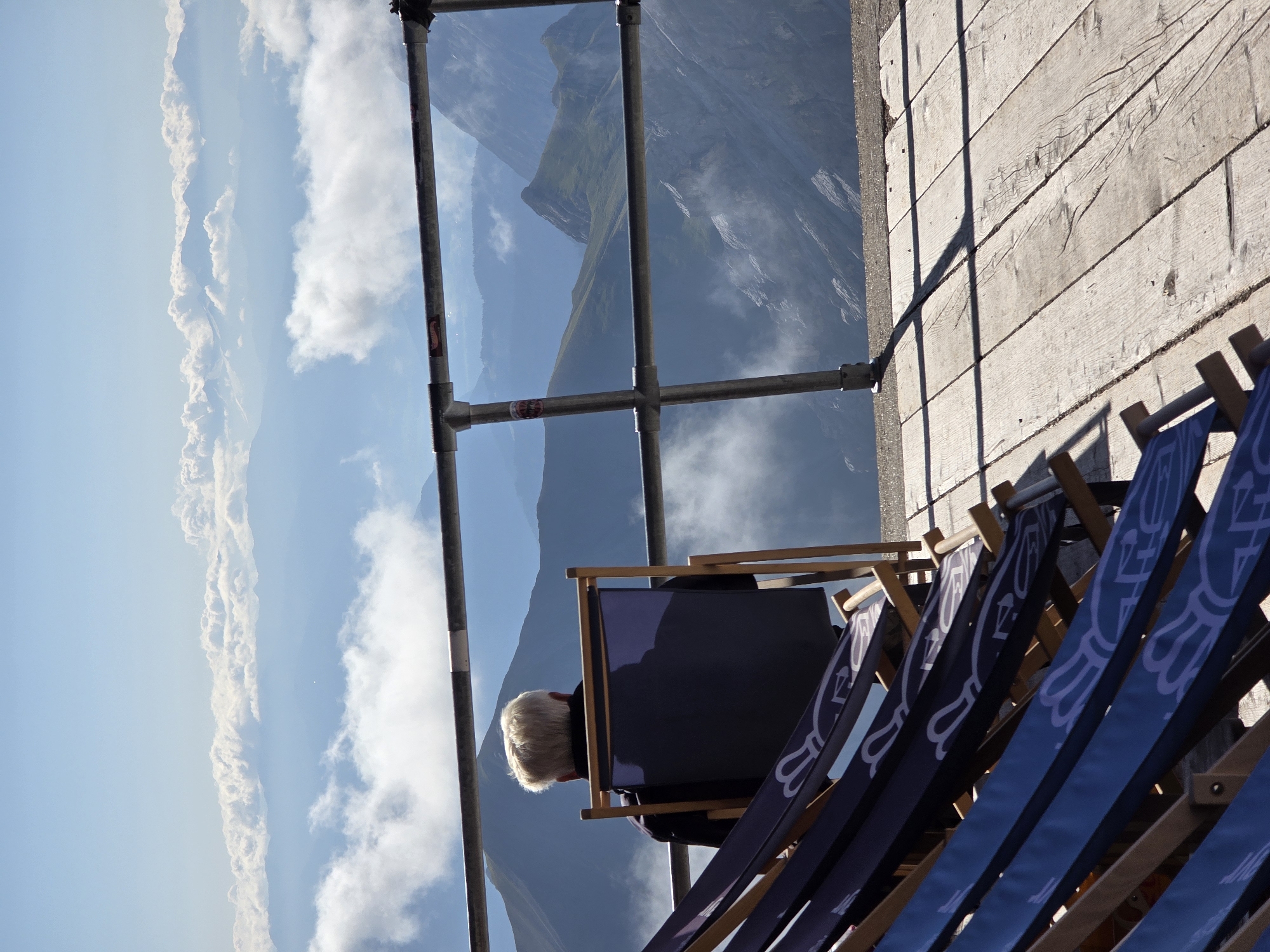 An elderly person with blonde hair sits in a dark blue deck chair facing away from the camera. The person is wearing a dark jacket. The deck chair is placed on a wooden platform, with several other identical deck chairs in the foreground, some of which are folded. A metal railing runs across the scene, separating the platform from a breathtaking view of a mountain range. The mountains are layered, with some peaks obscured by clouds. The sky is a clear blue with a few scattered clouds. The scene appears to be from a high vantage point, likely a tourist attraction offering panoramic views. This view is characteristic of the Zugspitze, Germany's highest mountain, located near Garmisch-Partenkirchen.