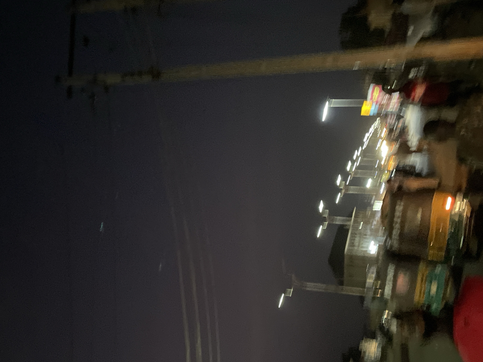 The image shows a nighttime street scene in what appears to be an urban area. Streetlights illuminate a road lined with buildings and businesses. There are people walking on the sidewalk, and vehicles including a three-wheeled vehicle (tuk-tuk or auto-rickshaw) are visible on the road. The location is identified as Bukuru, Nigeria. The scene appears busy, with indications of activity on both the street and sidewalk. Overhead power lines are visible, adding to the urban landscape.