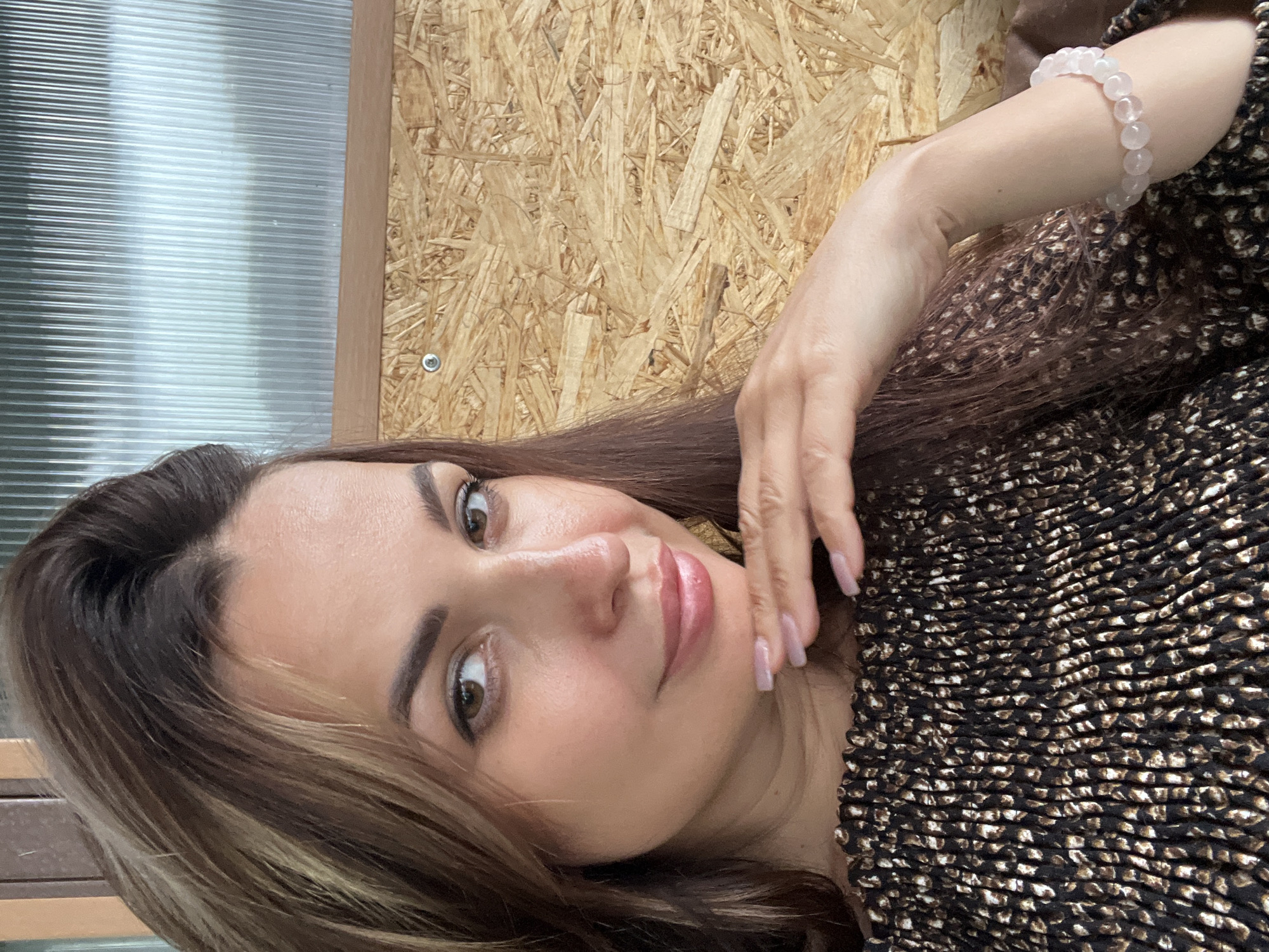 A woman with long, brown hair and brown eyes is taking a selfie. She is wearing a dark, off-the-shoulder top with a textured pattern. Her right hand is raised to her chin, and she is wearing a light-colored bracelet on her left wrist. The background appears to be outdoors, with a wall made of wood panels and a corrugated metal roof visible. The overall lighting suggests it is daytime. The location is not identifiable within the image.