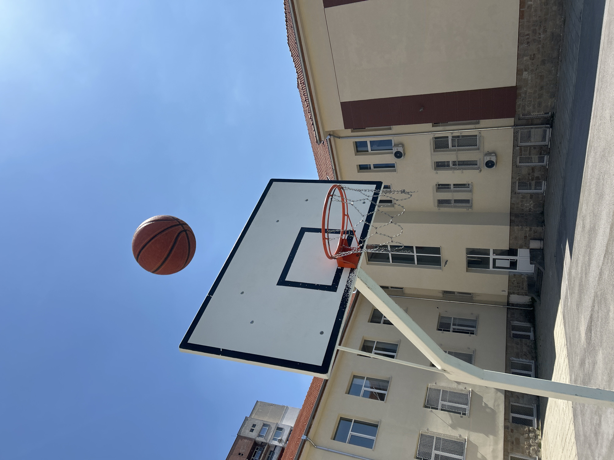 The image captures a basketball in mid-air, positioned to the left of a basketball hoop with a white backboard outlined in black. The hoop features a chain net. Behind the hoop and backboard, a multi-story building with light-colored walls and red-brown accents is visible. The building has multiple windows, some of which are secured with mesh coverings, and a few air conditioning units are mounted on the exterior. The sky is a clear, bright blue. Based on the architectural style and the use of Cyrillic characters potentially on signage not clearly visible, the location could be Sofia, Bulgaria. The image suggests an outdoor basketball court adjacent to an urban building.