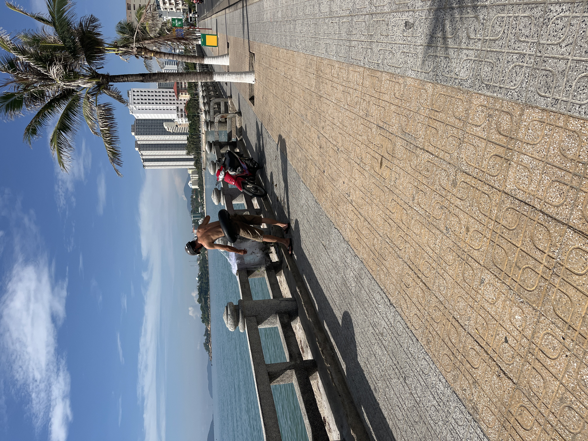 The image shows a man standing on a sidewalk next to a stone railing in Bac Nha Trang Ward, Vietnam. He is wearing shorts and a helmet, with no shirt. He appears to be using a fishing net near the water. Behind him is a red motorcycle parked on the sidewalk. In the background are palm trees, city buildings, and a bright blue sky with clouds. The sidewalk is tiled with alternating patterns of brown and gray stones. The water appears calm and stretches out to the horizon.