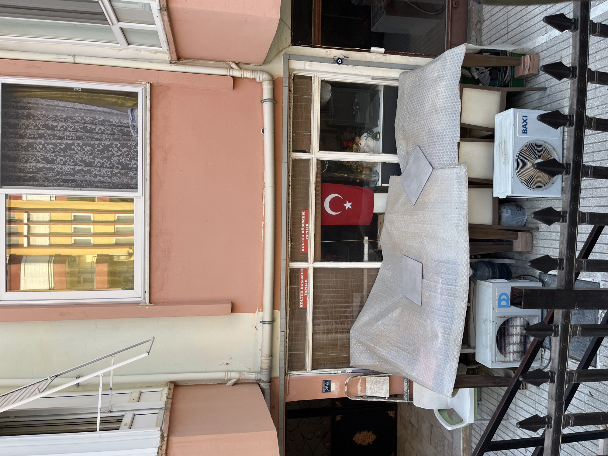 The image depicts the exterior of a building in Alanya, Turkey. The facade is painted in shades of pale pink and cream. On the second story, a window with white trim reveals yellow and orange buildings reflected in its glass, along with a decorative lace curtain. Below this, on the ground floor, is a shopfront with a large window displaying bamboo blinds. Two red signs with white lettering, reading "KOLTUK DÖŞEMESİ YAPILIR" (Upholstery is done), are visible on the shopfront window. A Turkish flag hangs inside the shop.
Outside the shop, several objects are present. Two white air conditioning units are placed on the ground. One unit, on the left, has a blue logo with white letters that appears to be "DD". The other unit, on the right, displays the brand name "BAXI". Behind the air conditioners, chairs and tables are covered with bubble wrap, suggesting they are either stored or protected. A large clear plastic water jug is also visible.
To the left of the shop entrance, a white stepladder leans against the wall. A sign with "28A" is affixed to the wall above the ladder. A dark metal fence with pointed finials runs along the bottom of the frame, partially obscuring the view of the ground level. The overall scene suggests a commercial establishment, likely an upholstery shop, with outdoor storage or display elements.
