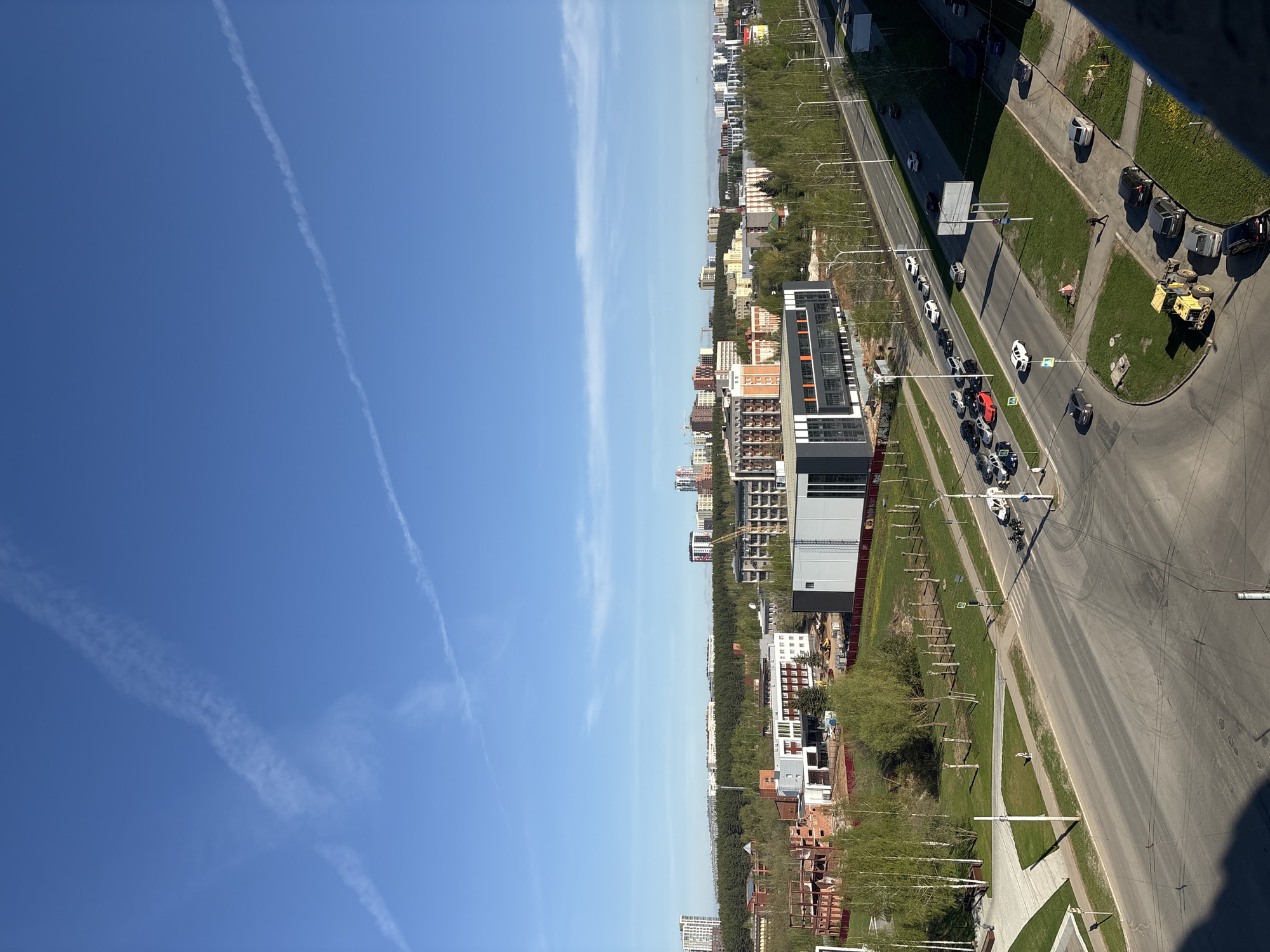 The image captures a high-angle view of a section of Izhevsk, Russia, on a bright day with a clear blue sky marked by contrails. The perspective is slightly rotated counter-clockwise. The urban landscape is divided into two main areas: the left side features the sky, while the right side showcases the city layout.
Buildings of varying heights and architectural styles populate the scene. A modern building with gray and black facade stands out. Further back, are residential apartment blocks. Patches of green spaces and trees are interspersed between the buildings.
A multilane road runs diagonally through the scene, lined with trees and streetlights. Cars, including a red one, are visible. A construction vehicle sits on a patch of grass. A pedestrian crossing is also visible.