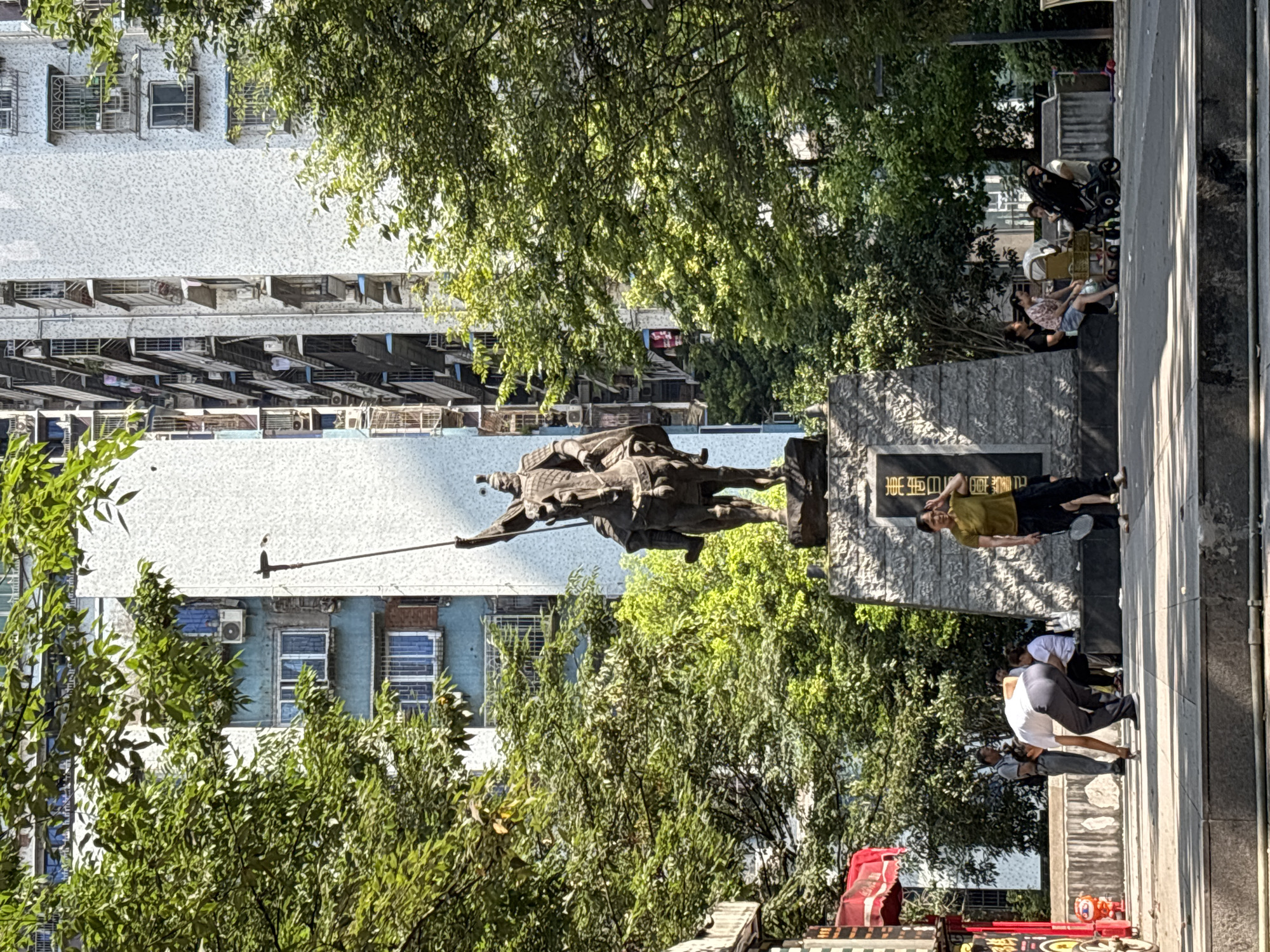 A statue of a mounted warrior, identified by Chinese inscriptions on the monument's base as being located in Yuzhong District, China, dominates the center of the frame. The statue is positioned in front of a tall, modern apartment building with a white, textured facade, and to the right of a multi-story building with blue accents. Lush green trees partially obscure the buildings and surround the statue. In the foreground, on a paved plaza, several people are engaged in various activities. A man in a white shirt and grey pants is bent over, appearing to pick something up from the ground. To his left, another man stands. To the right of the statue, a woman in a gold shirt and black pants holds a badminton racket and shields her eyes from the sun. Further to the right, a group of people are seated on a stone bench, some with a baby stroller. The overall scene depicts a public urban space with historical and modern elements, with people interacting within the environment on a sunny day.