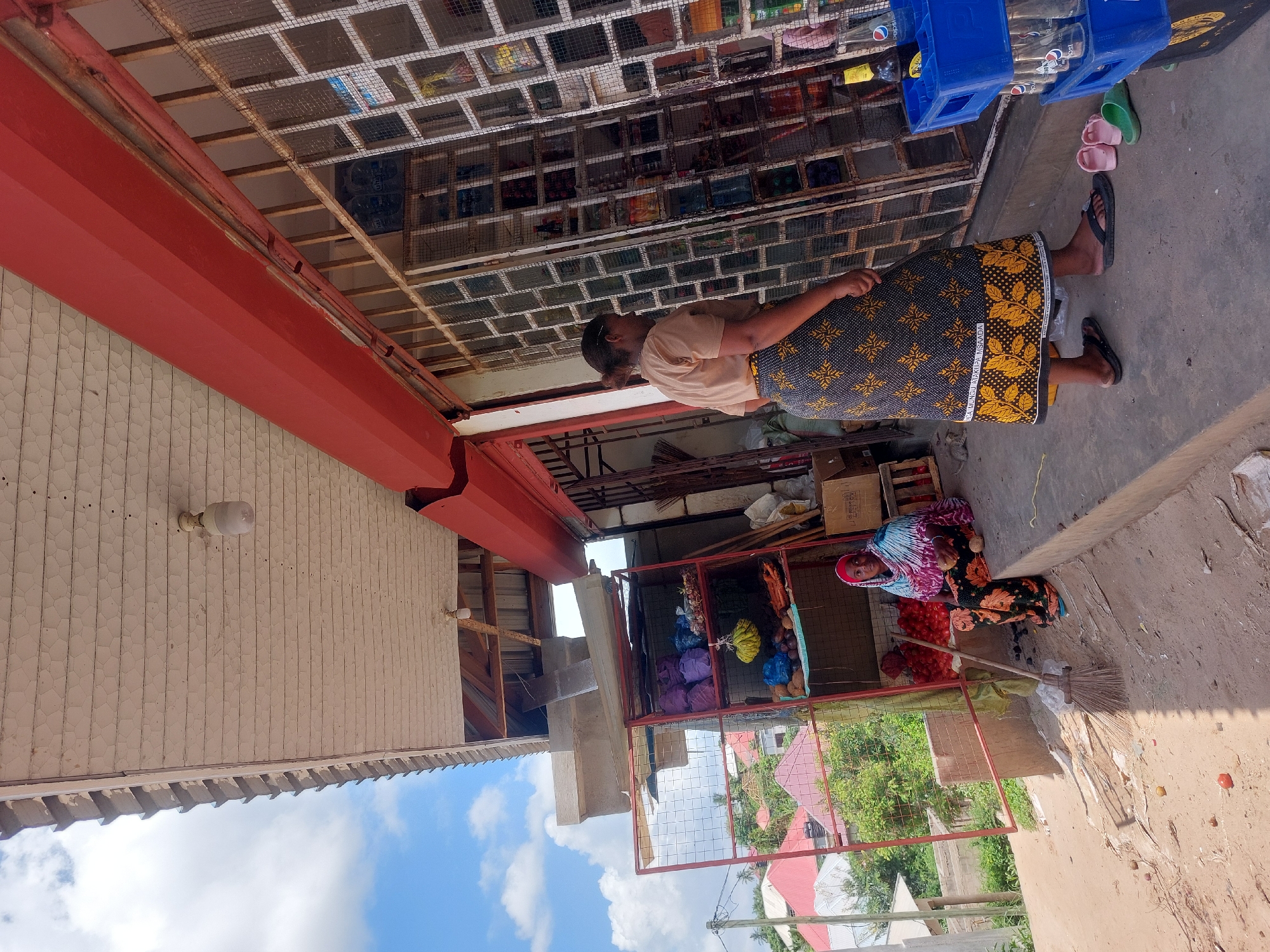The image depicts an outdoor scene in Dar es-Salaam, Tanzania. An African woman in a brown shirt and a yellow and dark-patterned skirt stands near a building. The building has a red overhang and white, textured walls with exposed light bulbs. Above the woman's head is a grid-like structure with various items placed within the squares. To the right, another African woman sits beside a metal-framed structure filled with fruits and vegetables. She wears a patterned headscarf, a floral shirt, and a long skirt. She sweeps the ground with a straw broom. Overhead, there's a collection of blue plastic crates, likely containing soda bottles. The surrounding environment includes partial views of nearby buildings, trees, and a bright blue sky with clouds.
