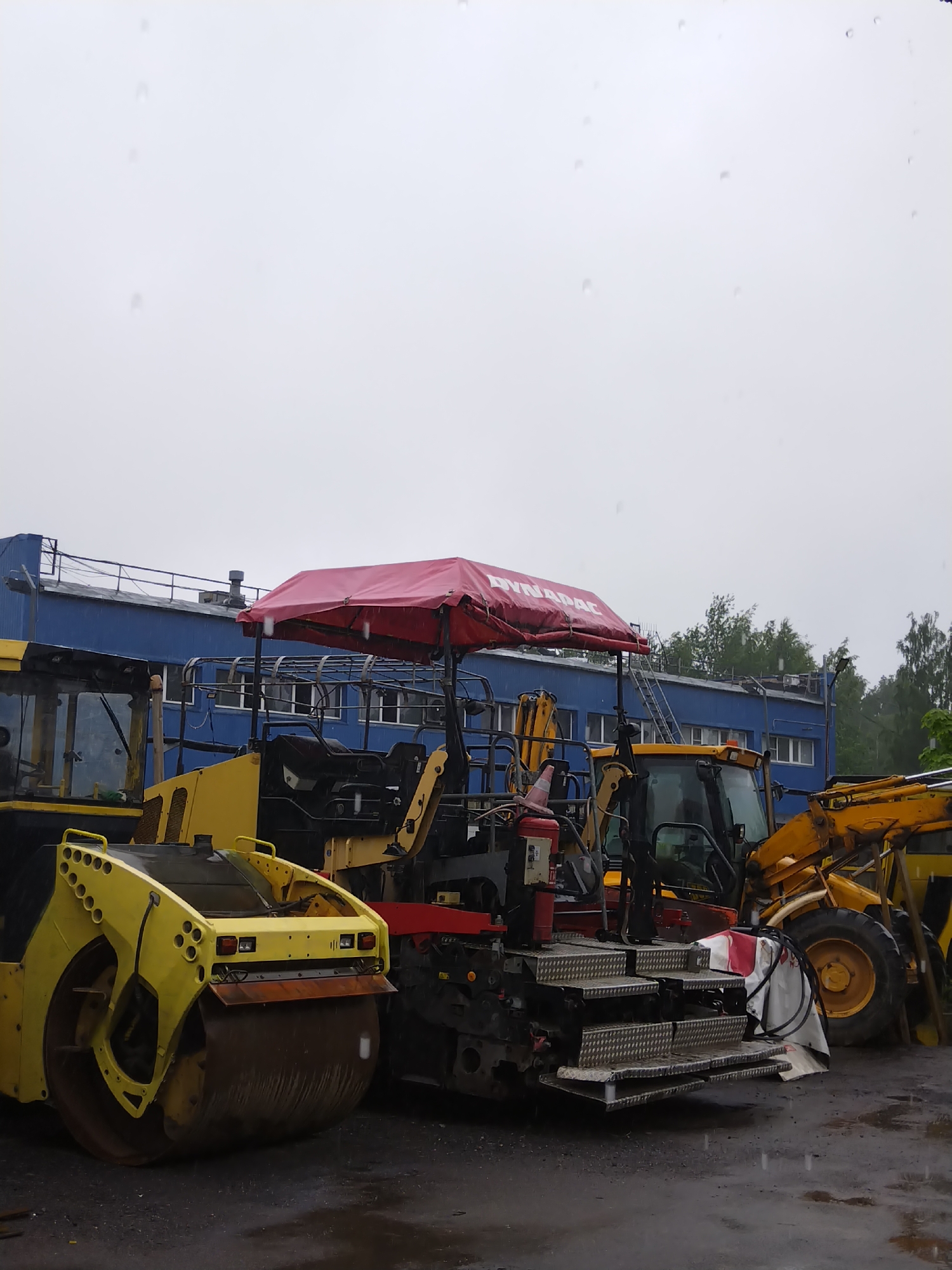 An outdoor, medium shot, eye-level, eye-level, vertical photograph shows construction equipment parked in front of a blue building. The building is a long, rectangular, two-story structure with a darker blue roof and a railing along the top. The building has many windows with dark frames and lights visible behind them. Trees are visible in the background behind the building. In the foreground, there is a bright yellow road roller with a large metal drum at the front. To the right of the road roller, there is a yellow asphalt paver with a red awning labeled "DYNAPAC" stretched over the operator's area. The paver has multiple metal steps leading up to the operator's seat, which is black. To the far right, part of a yellow backhoe loader is visible, with its front loader arm extended upwards. The ground is wet and appears to be asphalt, with puddles reflecting the overcast sky, indicating rain. Several raindrops are visible in the air and on the surfaces of the equipment. The scene appears to be a storage or operational yard for construction machinery. The location is the city of Pargolovo, Russia.