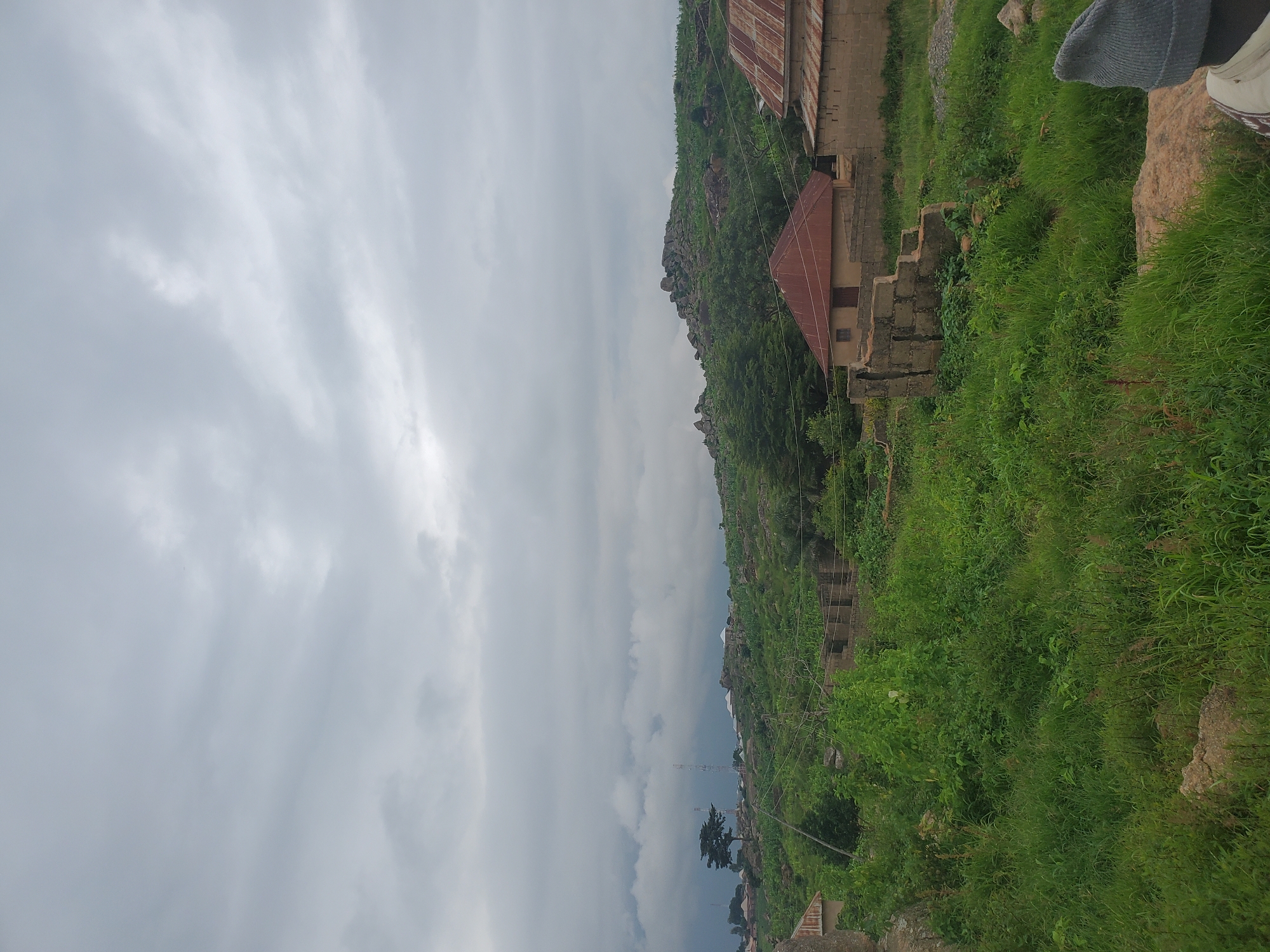 This image depicts a panoramic view of a semi-rural area, likely in Jos, Nigeria, under a cloudy sky. In the foreground, lush green vegetation dominates, with patches of dry grass and rocks. Partially constructed buildings, made of concrete blocks, are visible, suggesting ongoing development or unfinished projects. One building features a corrugated iron roof. Further back, a gently sloping hill is covered with dense greenery and scattered rock formations. Several telecommunication towers can be seen on the horizon, along with scattered houses with white roofs, indicative of a settlement. A single, mature tree stands out against the landscape. The overall atmosphere is overcast, suggesting impending rain or recent precipitation. A person wearing a grey beanie and part of a jacket is visible in the lower right corner, looking out at the scene. The scene appears to be a natural landscape interspersed with signs of human habitation and development.