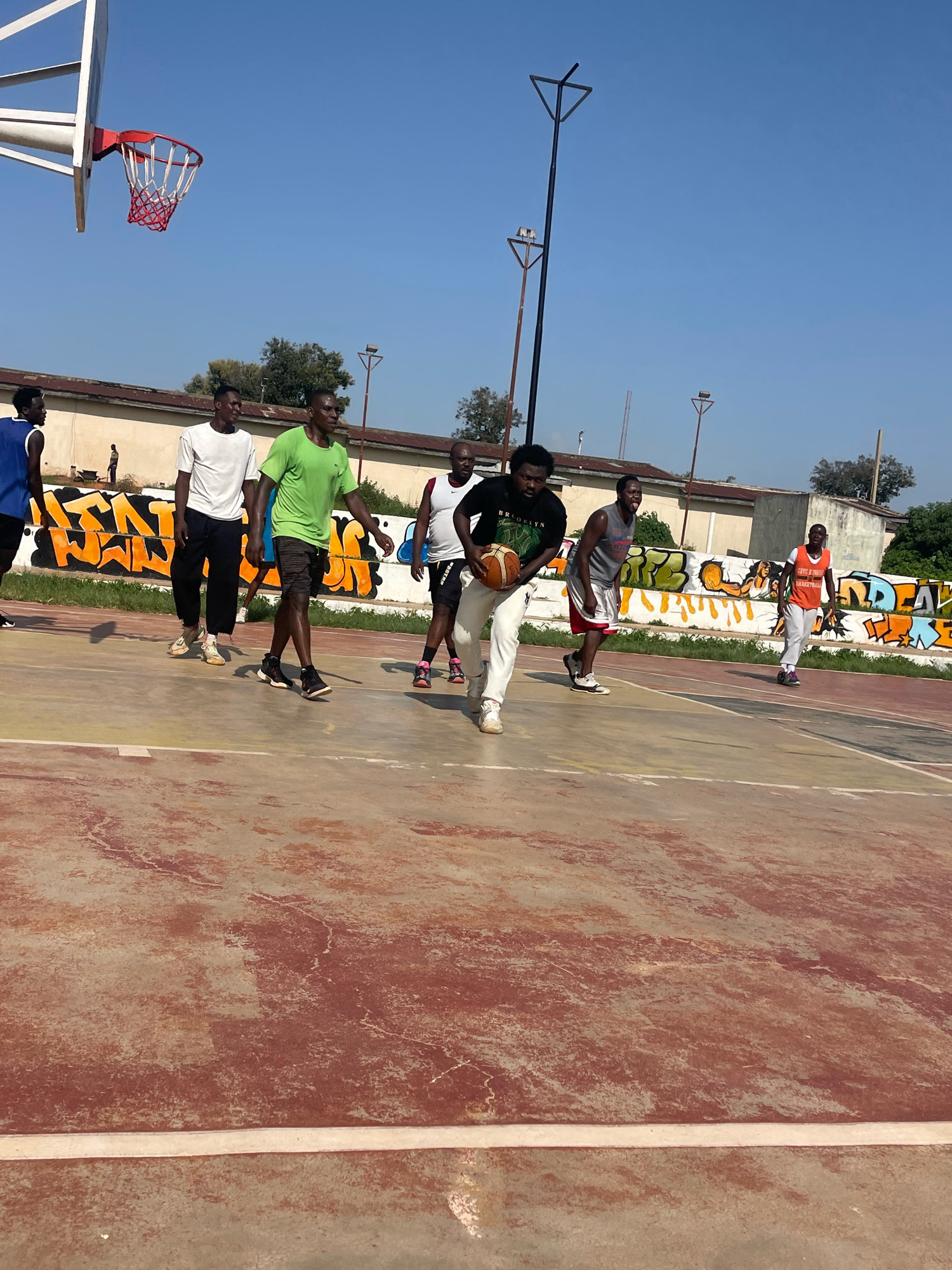 A group of men are playing basketball on an outdoor court under a clear blue sky. The court surface is red and cracked in places. In the background, there is a low building with graffiti on the wall. The players are dressed in athletic wear. One player in the foreground is holding a basketball. There is a basketball hoop in the upper left corner of the image. The scene appears to be in Jos, Nigeria, given the context provided.