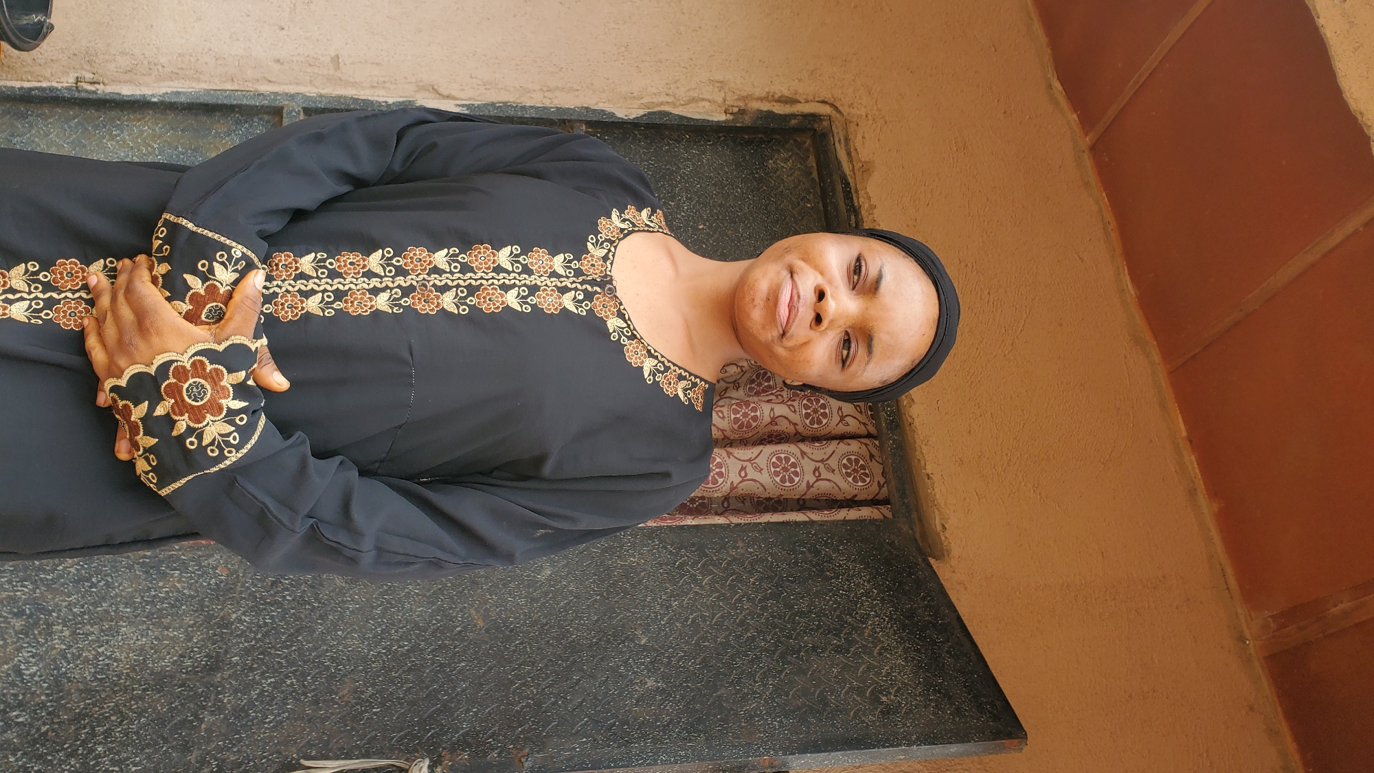 A medium shot captures a young woman wearing a black hijab and a black, long-sleeved dress with ornate gold and brown embroidery. The dress features a floral design along the neckline, down the center of the front, and on the cuffs. The woman has her hands clasped in front of her. She is looking slightly to her right with a neutral expression. She is standing outdoors, in front of a textured, beige-colored wall. To her right, there is a dark metal door with a patterned texture, and behind her, a patterned curtain is visible. Above her head, there is an overhang with a reddish-brown tiled roof. The lighting suggests it is daytime. The overall setting appears to be the exterior of a building, likely a residence or a small shop. The environment is consistent with an urban setting in Jalingo, Nigeria.