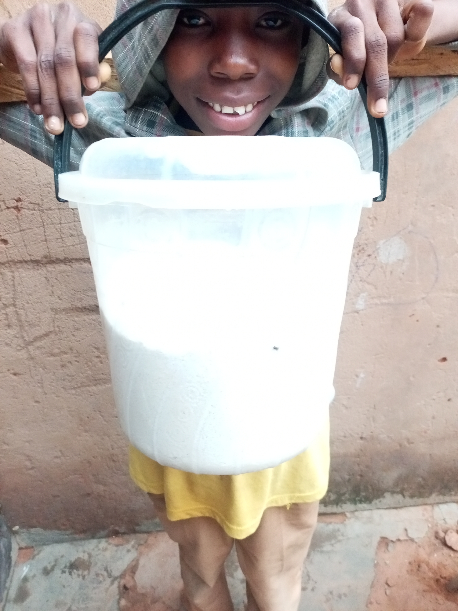 A young person, likely a child, is holding a large, white plastic bucket filled with a white substance. The person is wearing a hooded grey and white checkered shirt and a yellow garment underneath, with brown pants. They are looking at the camera and smiling, with their hands gripping the black handles of the bucket. The background appears to be a textured, reddish-brown wall, with a concrete floor showing some debris. The lighting suggests an outdoor setting, possibly in an urban environment like Gombe, Nigeria, given the wall's texture and the general aesthetic. The focus is on the person and the bucket they are holding.