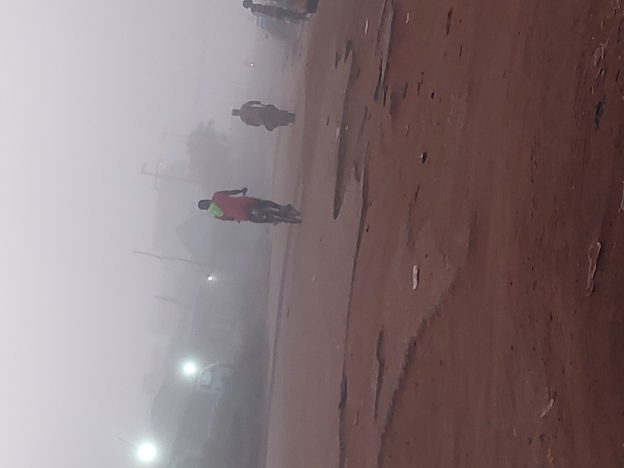 A foggy, overcast scene unfolds on a reddish-brown dirt road, suggesting a location like Bukuru, Nigeria. The poor visibility indicates either early morning or late evening conditions, possibly during a Harmattan season. In the foreground, the road is marked by numerous potholes and debris scattered across its surface.
Three individuals are visible. The closest subject, positioned in the mid-left of the frame, is on a bicycle, facing away from the camera. They are wearing a red shirt with a yellow and green sash or garment draped over their left shoulder. Further back, a second figure, also on a bicycle, is almost obscured by the fog, appearing as a dark silhouette. A third individual, standing, is visible on the right side of the frame, also partially obscured.
In the background, streetlights pierce through the fog, illuminating some structures and vehicles. The presence of these lights suggests a settlement or urban area. The overall atmosphere is one of muted light and limited visibility, characteristic of foggy conditions.