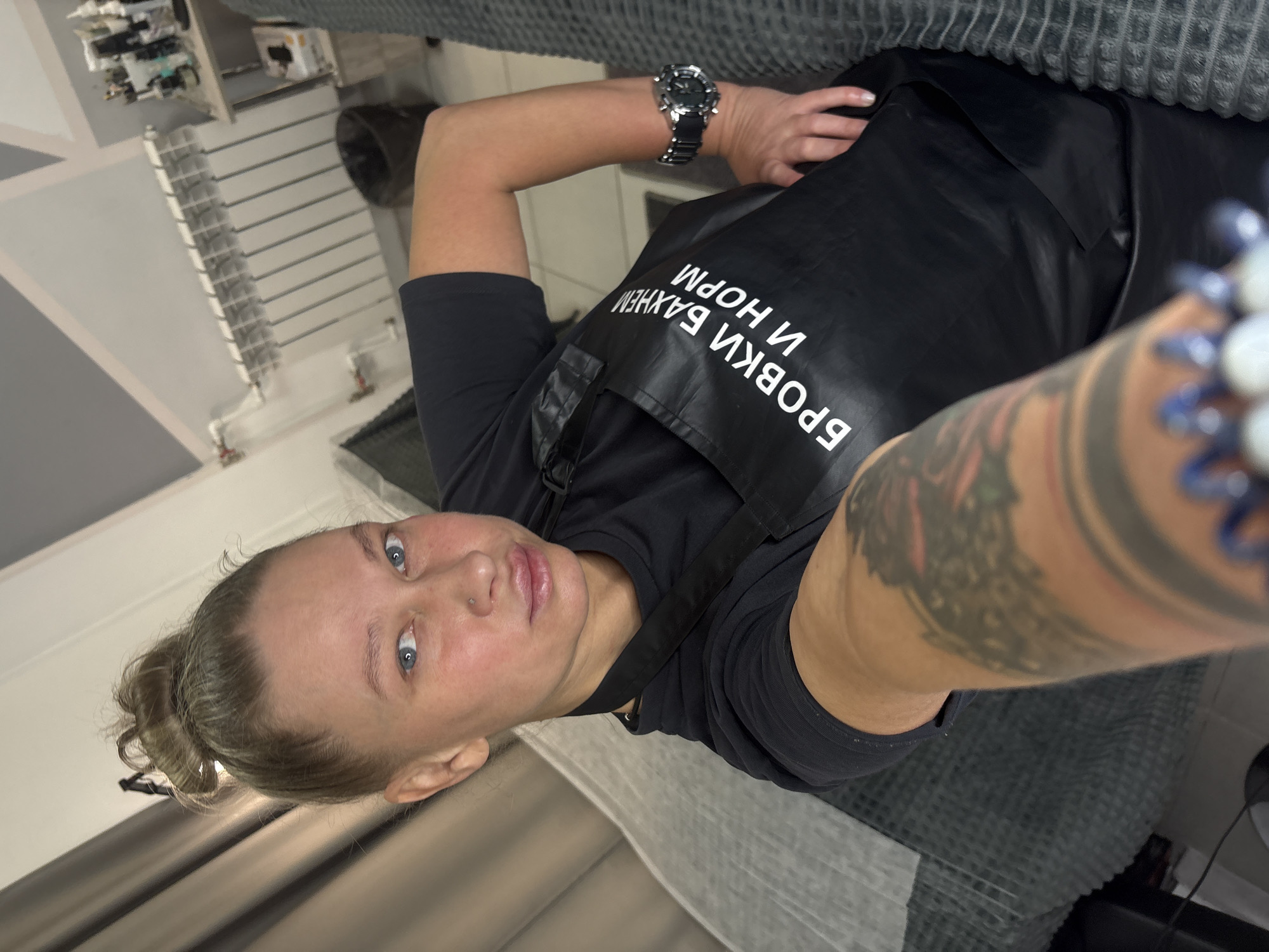 This is a selfie-style portrait of a woman taken indoors, likely within a beauty salon or studio in Perm, Russia. The woman, with light blue eyes and a nose piercing, looks directly into the camera with a neutral to slightly serious expression. Her light brown hair is tied back in a bun.
She is wearing a short-sleeved black top and a black apron. The apron features white Cyrillic text that reads "БРОВКИ ВАЖНЕЙ И НОРМ," which translates to "Brows are more important and norm" or "Brows are more important and okay." This text strongly suggests her profession is related to eyebrow artistry or beauty services. She wears a silver watch on her left wrist and a large, colorful tattoo is visible on her right forearm, along with a blurred, multi-beaded bracelet.
The background shows elements of a professional workspace. To the left, there are shelves with various small items, possibly beauty products or tools, and a white fixture resembling a radiator or a shelving unit with horizontal slats. A textured grey surface, likely a treatment bed or couch, is visible behind and above her, spanning the top and right side of the image. The setting appears clean and well-lit by indoor lighting.