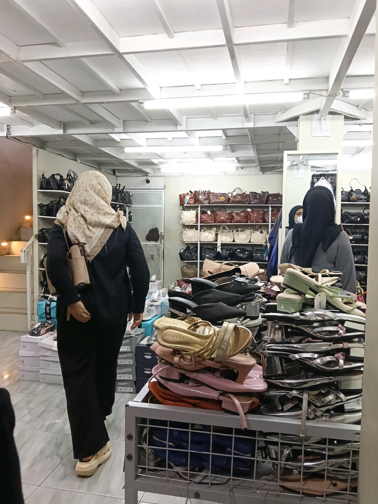 The image shows the interior of a shoe store located in Dengok, Indonesia. In the foreground, a person wearing a hijab and dark clothing is walking away from the camera, carrying a small purse. They are wearing light-colored clogs. In the background, shelves are stocked with various handbags. On the right side of the image, another person wearing a black hijab and a grey sweater is visible, along with a display of women's shoes. The store appears to be well-lit, with fluorescent lighting fixtures on the ceiling. The floor is tiled.