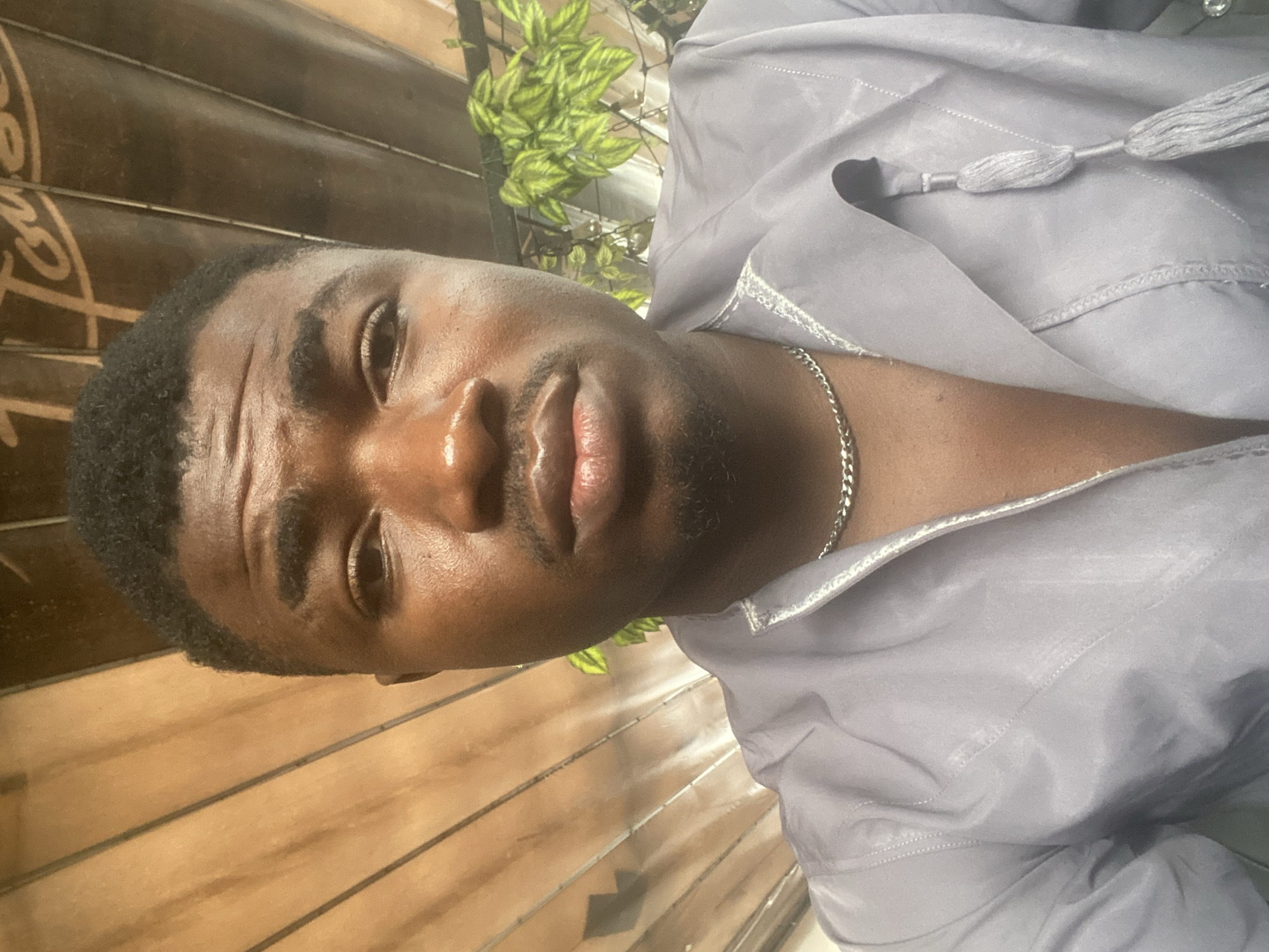 A close-up, eye-level selfie depicts a young Black male from the chest up, positioned against a varied background. He has short, dark, curly hair, prominent eyebrows, and light stubble on his chin and upper lip. His forehead and nose show a slight sheen. He wears a light grey or lavender-colored long-sleeved shirt with an open collar, featuring light-colored trim and a decorative tassel detail on the right side. A silver-colored cuban link chain necklace is visible around his neck. The man maintains a neutral expression, looking directly at the camera. To the left of the subject, the background consists of diagonally oriented light brown wooden slats or panels. To the right, a darker brown surface, possibly a ceiling or awning, displays partial script lettering in a lighter color, appearing to read "Tou..." or "Touse...". Below the lettering and to the subject's right, vibrant green leafy plants are visible. The scene is located in the city of Howayi, Nigeria.
