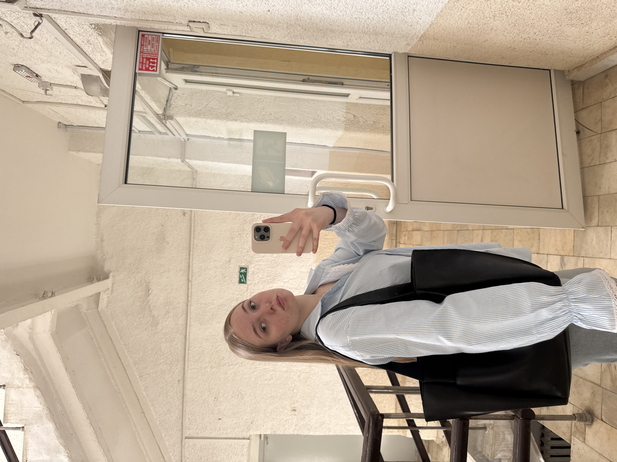 A young woman stands in what appears to be a stairwell in Perm, Russia, taking a selfie. The environment consists of white textured walls and stairs with metal railings. The foreground features the woman holding a smartphone; she is wearing a light blue and white striped blouse with ruffled cuffs and a black shoulder bag. Behind her, a closed white-framed door is visible with a rectangular window at the top. A fire safety sign in Russian is affixed to the upper left corner of the door. The surrounding stairwell seems to have rough, textured walls.