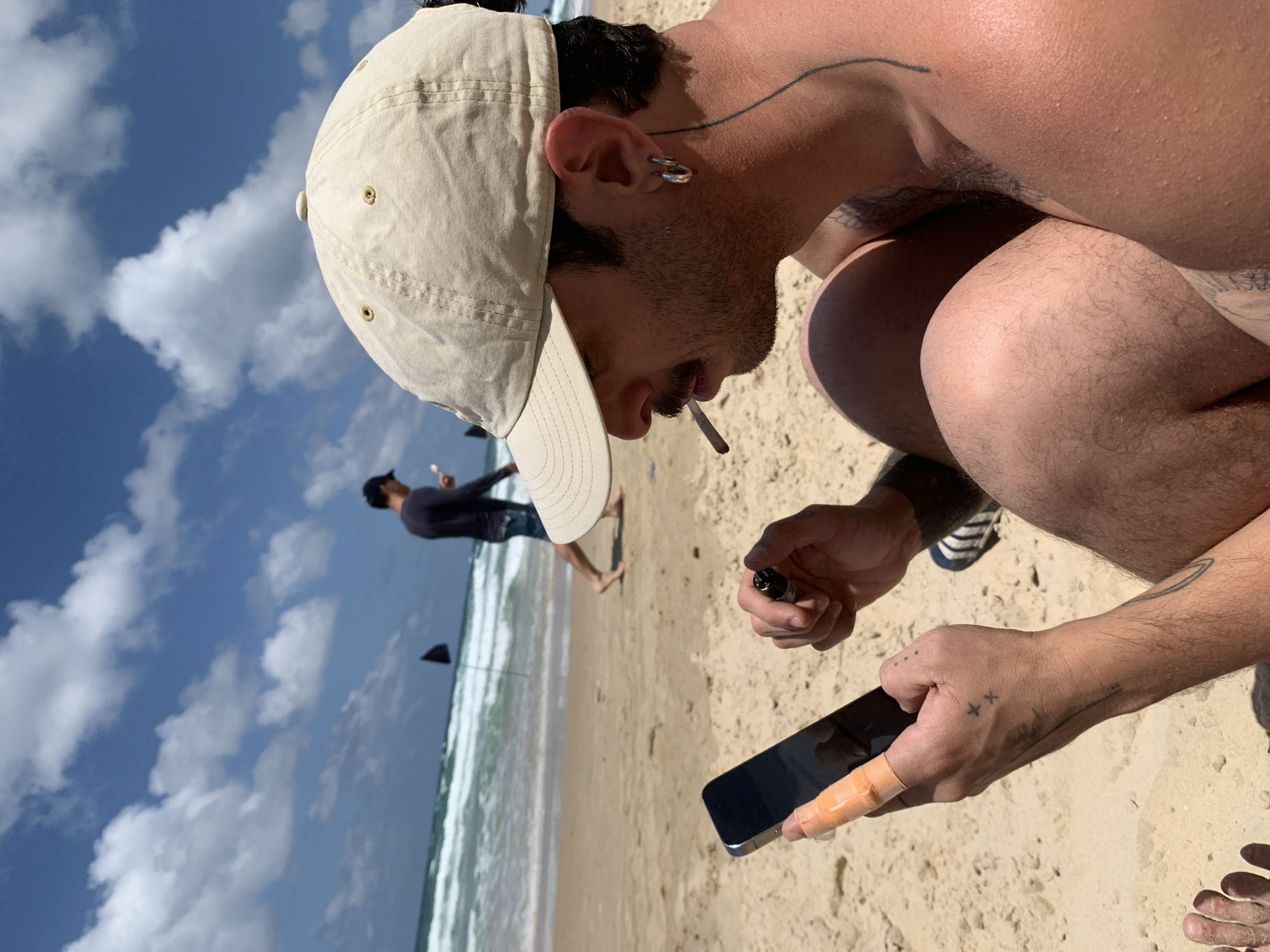 The photo depicts a man on a sandy beach, possibly smoking a cigarette or joint. He is wearing a tan baseball cap and has a silver earring. He is shirtless, with a visible tattoo on his upper arm and another on his neck. He is holding a black smartphone in one hand and what appears to be a vape pen or similar device in the other. There is a bandage on one of his fingers. In the background, another individual is walking along the shoreline. The sky is blue with scattered clouds. This is likely taken at a beach in Tel-Aviv, Israel, but there is no definitive indicator of location.