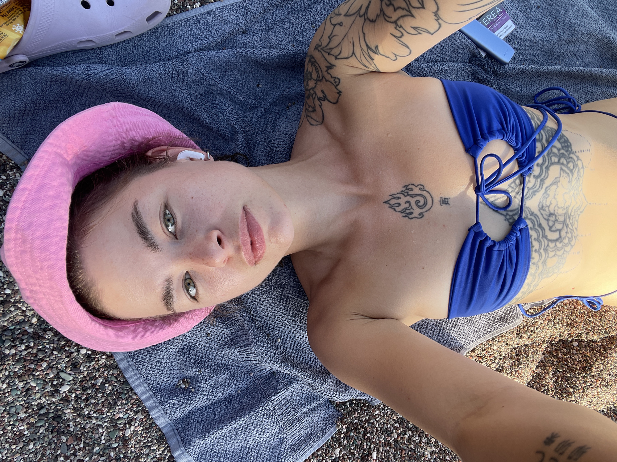 A young woman with tattoos is lying on a gray towel on a beach. She is wearing a blue bikini and a pink bucket hat. She has fair skin, green eyes, and light brown hair. She is lying on her back, looking up at the camera. An earpiece is visible in her left ear. Next to her is a pair of pink Crocs and a blue vape pen. The beach is covered in small pebbles. The location is likely Rafailovići, Montenegro, given the environment of a pebble beach.