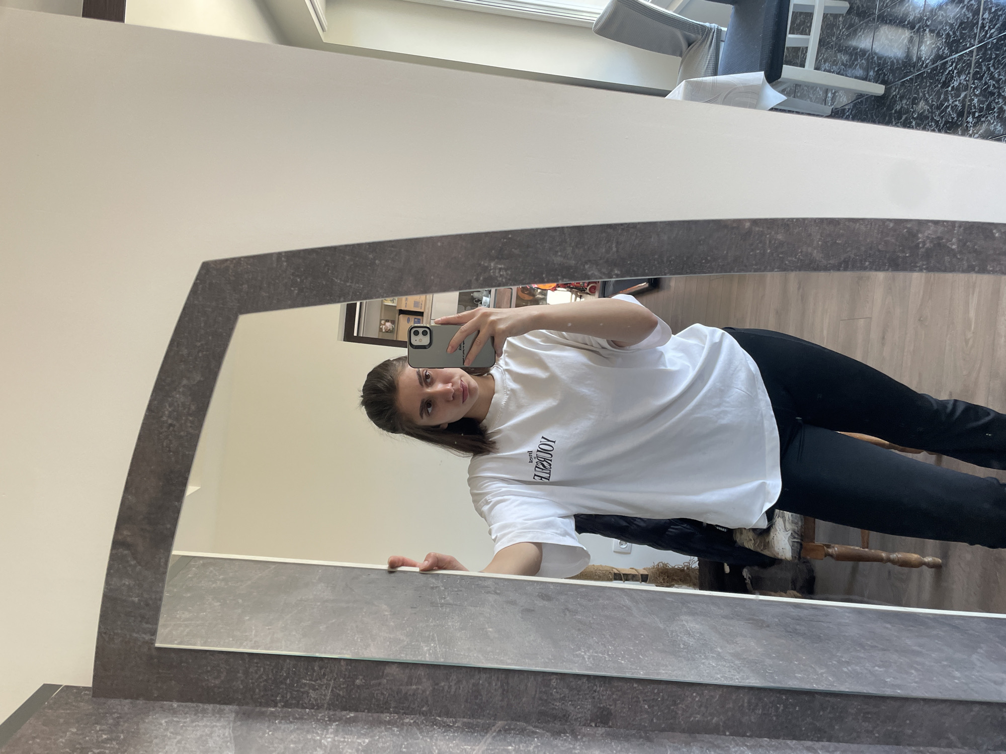 A young woman is taking a selfie in a mirror. She is wearing a white T-shirt and black pants. The mirror is framed with a gray, faux-marble material. The background shows a room with light-colored walls and a wooden floor. A chair and a window are visible in the reflection. It is impossible to determine the specific location in Sofia, Bulgaria from this image alone.