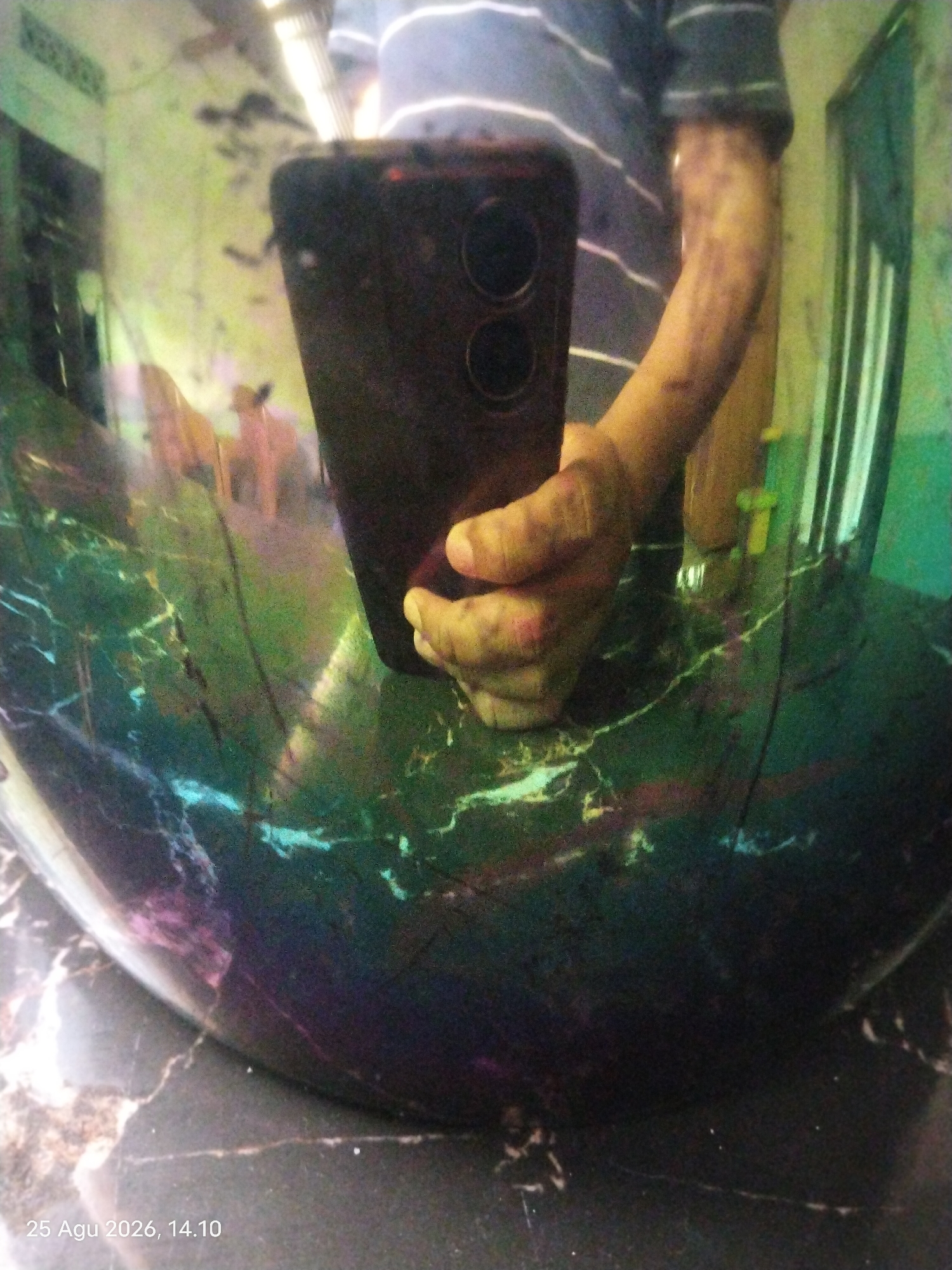 The image shows a reflection of a person holding a smartphone in what appears to be a highly reflective, possibly polished, dark surface with green and purple marbling. The person is wearing a horizontally striped t-shirt. The background of the reflection shows a room with vertical elements, possibly windows or blinds, and some indistinct figures. The date stamp on the image indicates "25 Agu 2026, 14.10", which translates to August 25, 2026, at 2:10 PM. The location is stated as Singkawang, Indonesia. The overall scene is abstract and distorted due to the reflective surface.