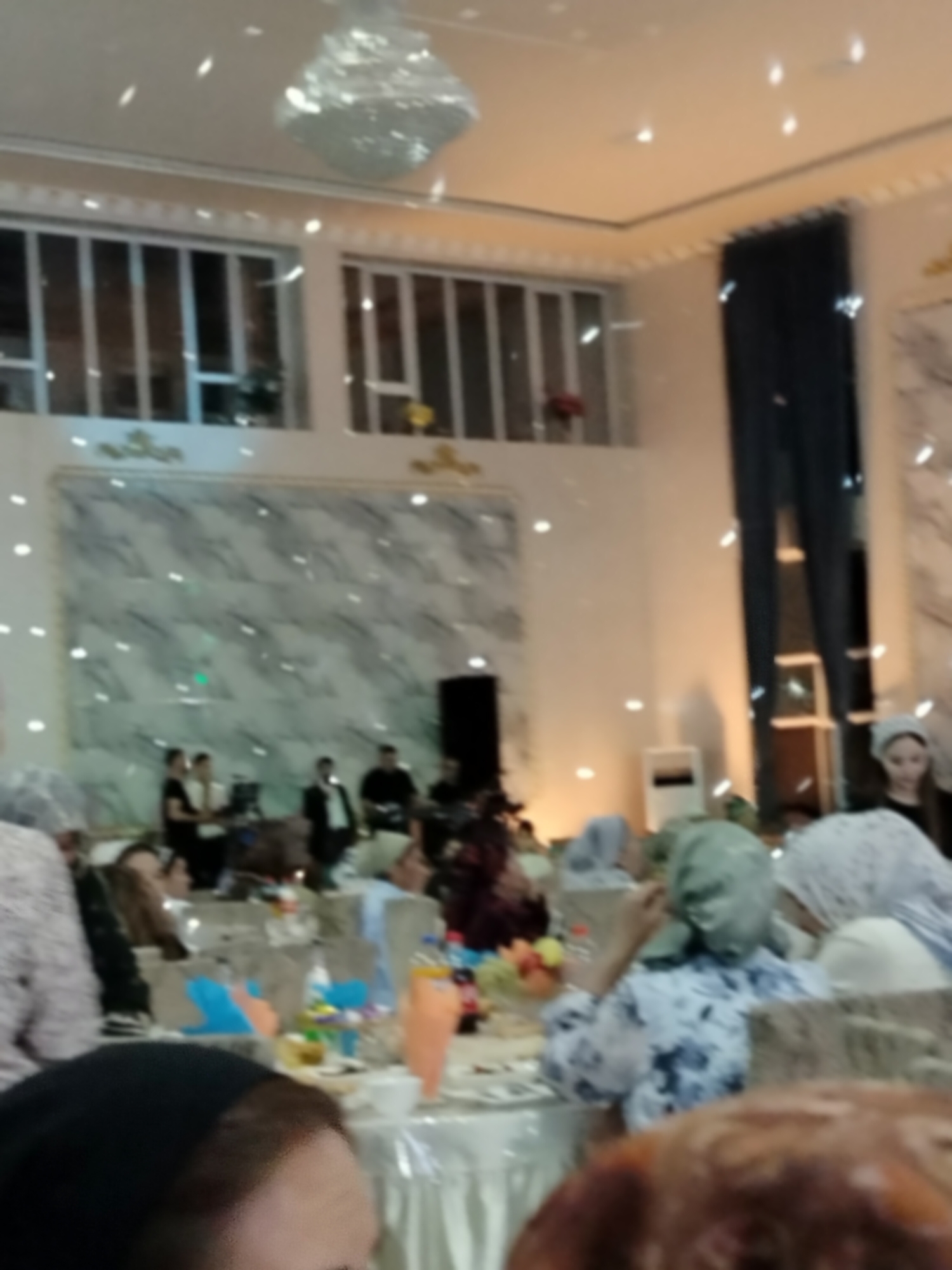 The image depicts an indoor event, possibly a wedding reception or banquet, taking place in a dimly lit hall with a white, draped ceiling. A large, multi-tiered chandelier hangs prominently from the center of the ceiling, reflecting numerous small, bright lights across the scene. The walls are adorned with large, marbled panels, and decorative window frames are visible in the background.
In the foreground, several people are seated at a long table draped with a white tablecloth. The table is set with various items, including plates, cups, and decorative elements like small baskets with colorful items and what appear to be fruit arrangements. The lighting creates a scattered, bokeh effect with numerous small, white circles of light.
Further back, a group of people are standing, some of whom appear to be engaged in conversation or preparing for the event. One individual in the background is wearing a white head covering and a dark garment. Another person is seen wearing a light-colored head covering and a patterned dress. The overall atmosphere suggests a gathering or celebration. The location is identified as Ақсуабат, Kazakhstan.