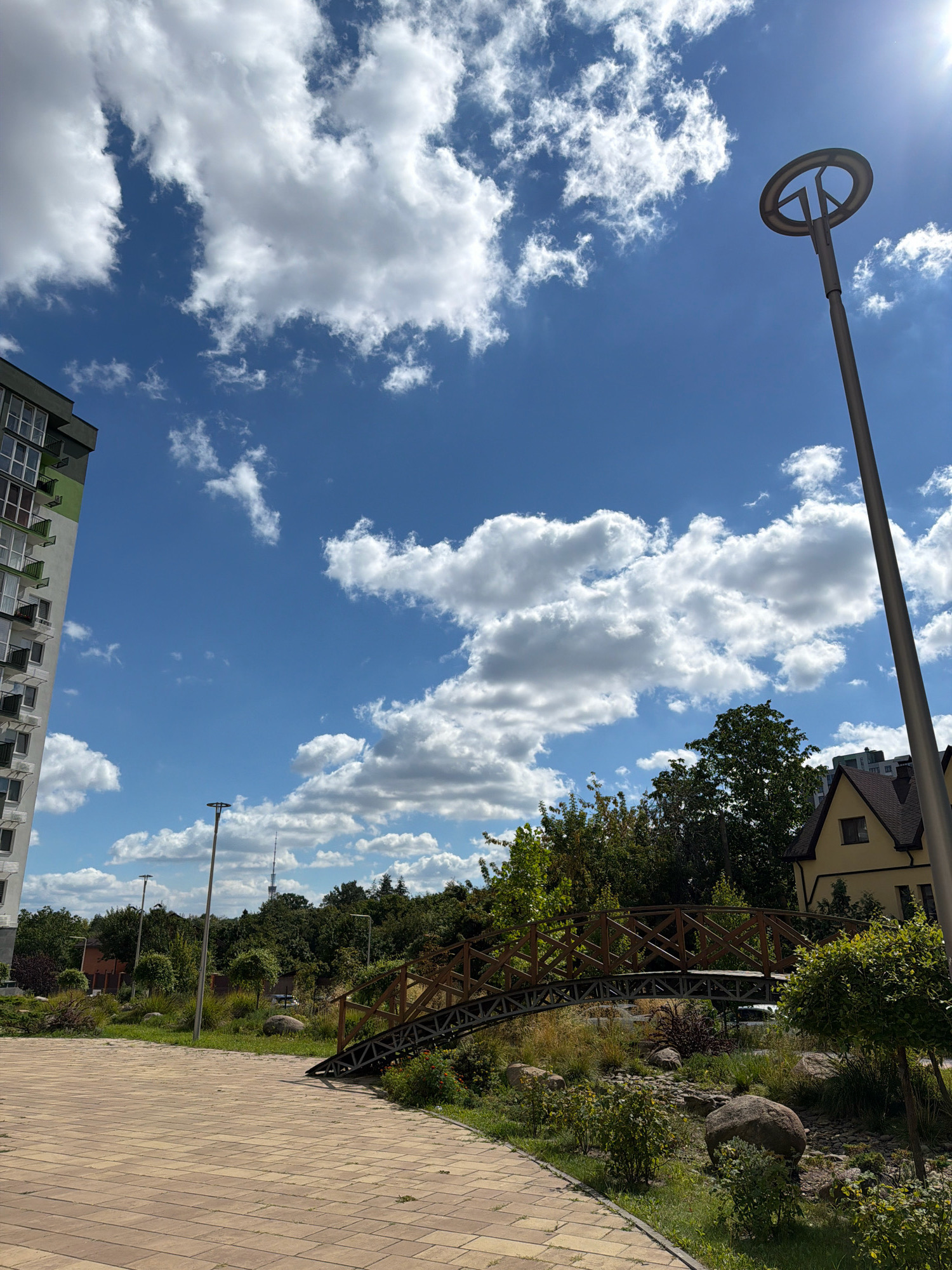 The image depicts an outdoor scene on a sunny day with a clear blue sky filled with fluffy white clouds. In the foreground, a beige brick pathway curves to the left. To the left of the pathway is a tall, modern apartment building with green and white accents and numerous balconies. Further into the scene, a small, arched wooden bridge spans a dry creek bed or decorative water feature with rocks and various plants. Behind the bridge, a cluster of green trees forms a natural backdrop, and a yellow house with a dark roof is visible among the trees. A tall, grey lamppost with a circular fixture stands to the right of the frame, and another lamppost is visible in the distance. In the far background, a tall tower, possibly a television or radio tower, can be seen rising above the trees. The overall environment appears to be a developed urban or suburban area with a park-like setting.