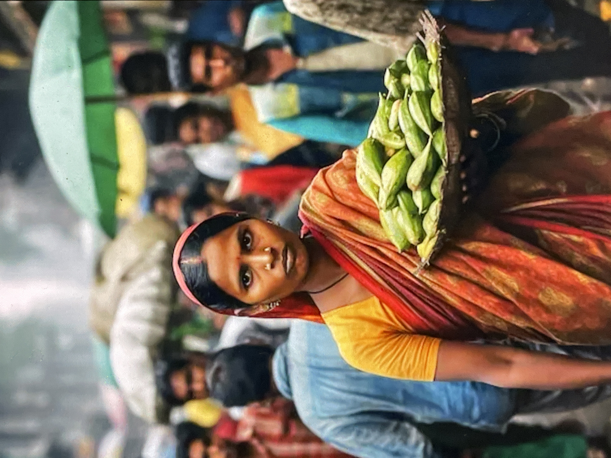 A woman in a vibrant orange and red sari stands in a crowded street, holding a basket of green vegetables on her head. Her dark hair is pulled back, and she wears earrings and a nose ring. Her expression is direct and unsmiling. Behind her, people mill about, some carrying umbrellas, suggesting a bustling marketplace. The background is blurred, but hints of buildings and other vendors can be seen. The image captures a moment of everyday life in what appears to be a busy urban environment, likely a market in Yalga, Russia. The overall impression is one of vibrant color and human activity.