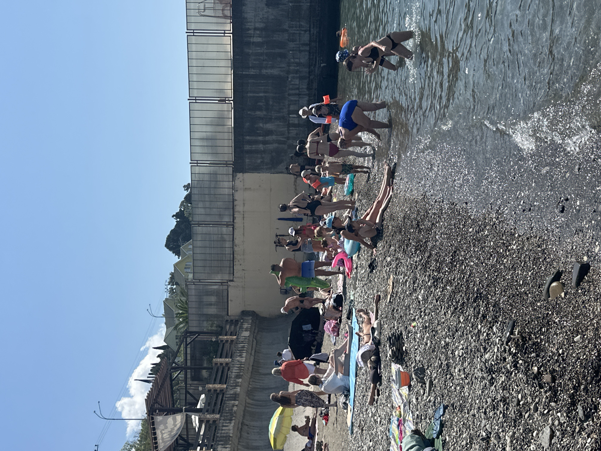The image depicts a crowded pebble beach on a sunny day, likely in Sochi, Russia, given the architecture and vegetation. In the foreground, people are sunbathing on towels and in swimwear on the dark grey pebbles. Some are lying down, while others are sitting or standing. The water is clear and blue, with several individuals wading or standing in the shallow areas near the shore.
Further back on the beach, more people are gathered. Some are under umbrellas, seeking shade. There are inflatable toys, such as a pink ring and a blue ball, scattered on the sand. A yellow umbrella provides shade for a couple of people. Beach bags and other belongings are visible.
Behind the beach, a concrete wall and fence line the area, with a structure that appears to be a part of a resort or public building, including a wooden-topped canopy. The background shows lush green trees and some houses on a hillside, characteristic of the coastal landscape of Sochi. The sky is a clear, bright blue with a few wispy clouds. The overall scene conveys a lively summer atmosphere at a popular beach destination.