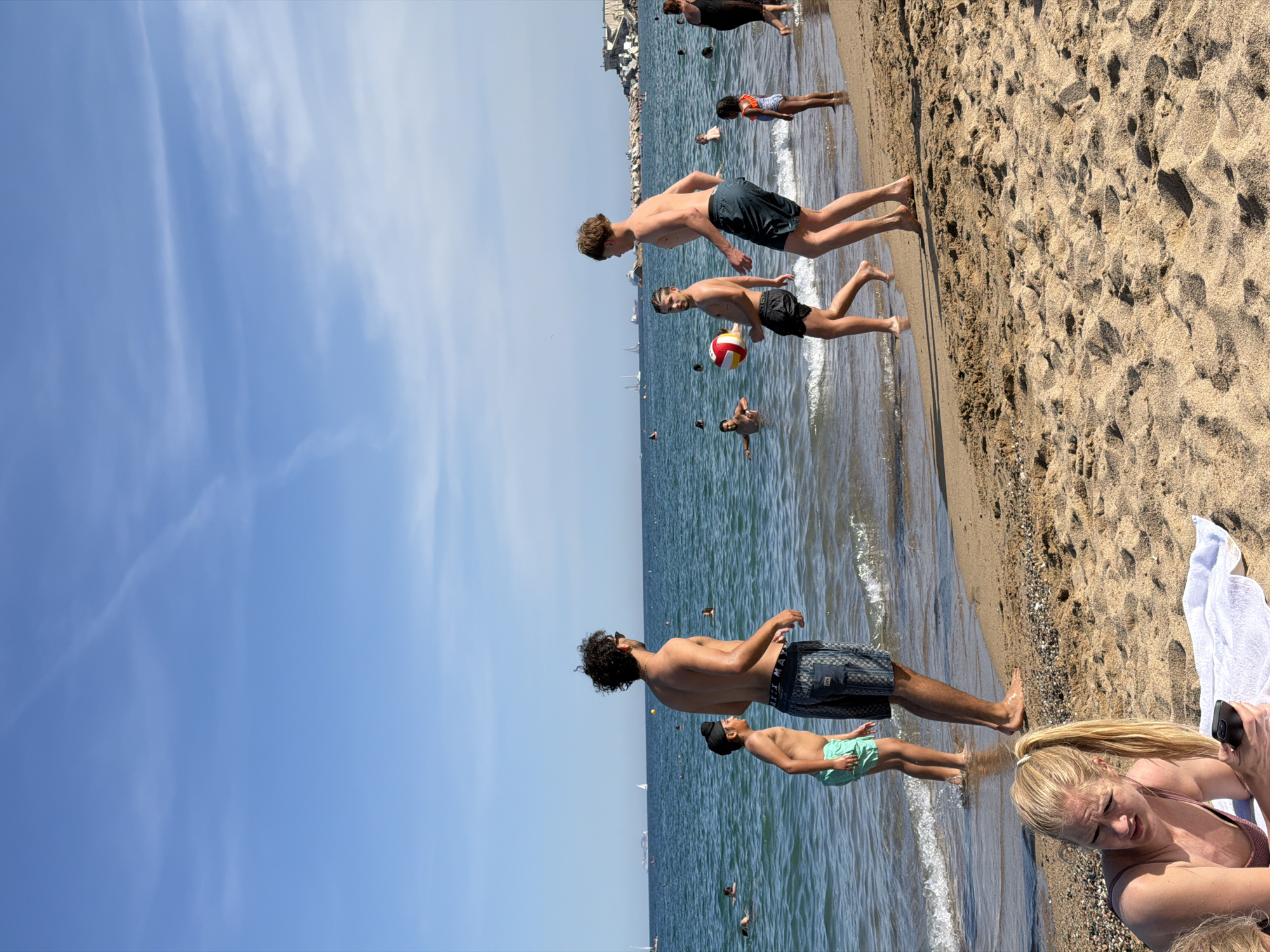 The image depicts a sunny day at Barceloneta Beach in Barcelona, Spain. The foreground features a woman lying on a towel on the sandy beach, looking at her phone. In the mid-ground, several people are engaged in recreational activities in the shallow water. Two young men are playing with a beach ball, one holding it and the other reaching out. Another young boy in green shorts stands near the water's edge. Further out in the sea, other individuals are swimming and enjoying the water. In the background, sailboats and a large cruise ship are visible on the horizon, with a breakwater and part of the city's architecture visible on the right side of the image. The sky is clear blue with some wispy clouds, suggesting good weather.