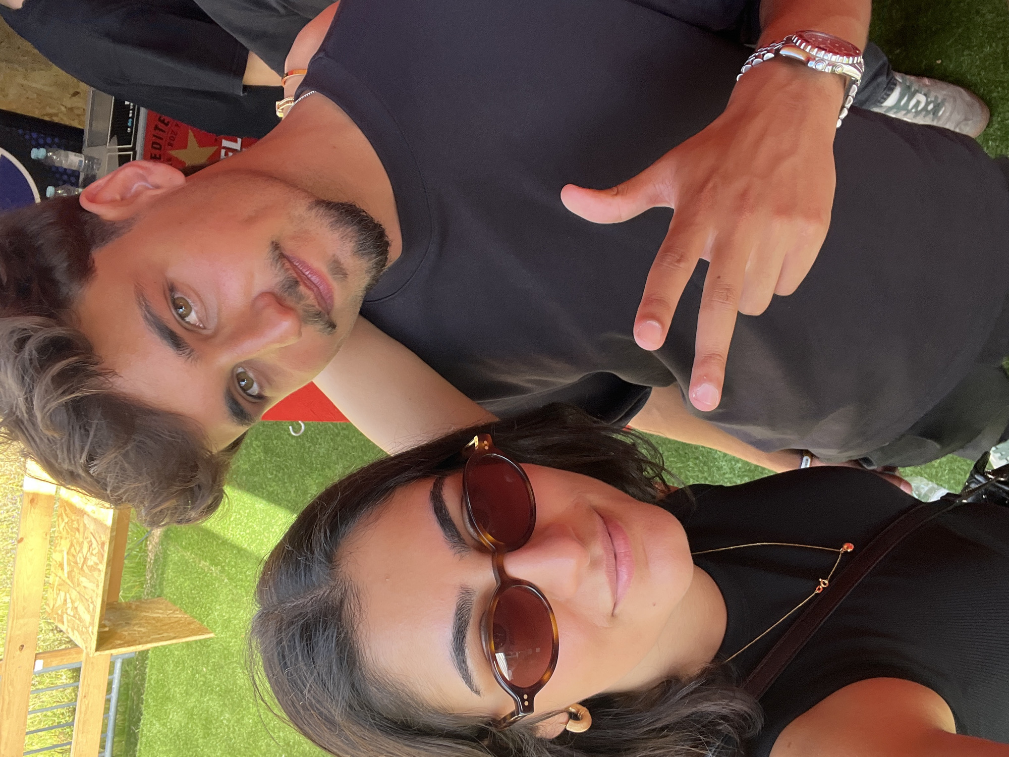 The image shows a young man and woman posing for a selfie outdoors. The woman in the foreground is wearing a black tank top, sunglasses, and a gold necklace with a red charm. She has dark hair and is wearing hoop earrings. The man behind her is wearing a black t-shirt and a silver watch. He has short dark hair and a goatee. He is making a peace sign with his right hand. The background shows artificial turf, a wooden structure, and some fencing. The overall scene suggests a casual outdoor event or gathering. The location is the city of Porto, Portugal.
