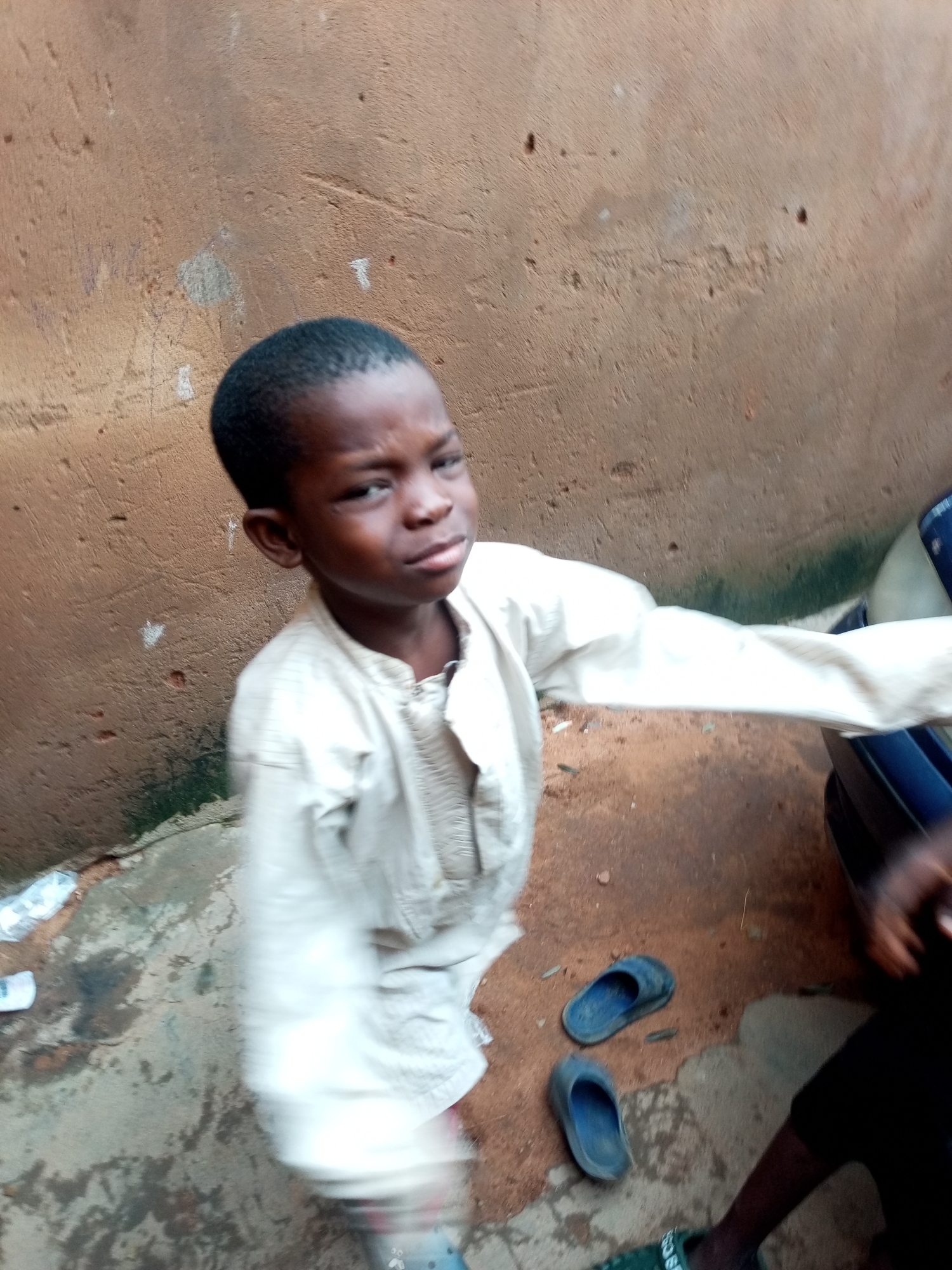 A young boy wearing a light-colored, long-sleeved tunic stands near a textured, brown wall. He is looking towards the camera with a slightly furrowed brow. The ground is uneven and appears to be a mix of dirt and concrete. Two blue flip-flops are visible on the ground to his right. A portion of a dark vehicle is visible on the far right of the frame, and some white plastic is scattered on the ground to the left. The overall environment suggests an outdoor setting, possibly a yard or alleyway in Tempure, Nigeria.