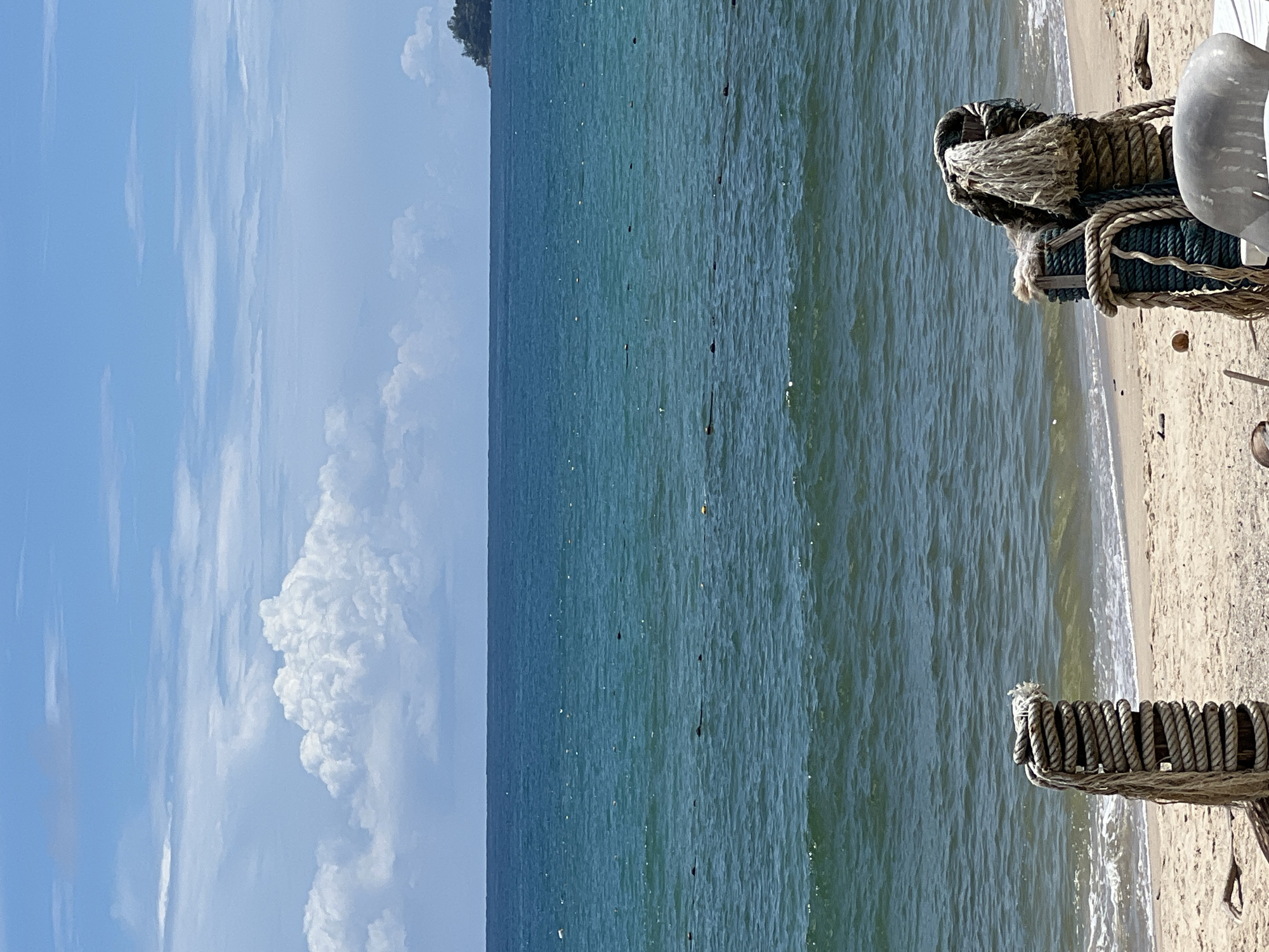 The image shows a beach scene with the ocean and sky. The foreground is dominated by two posts wrapped in thick ropes, suggesting a mooring area or a decorative beach element. The sand of the beach is visible at the bottom of the frame, with some small debris scattered on it. The ocean is a deep blue-green, with gentle waves rolling towards the shore. A line of buoys or markers can be seen in the water, possibly delineating a swimming area or a channel. The horizon separates the ocean from the sky, which is a bright blue with scattered white clouds. The clouds are fluffy and voluminous, with a large, prominent cloud formation in the upper left portion of the image. In the far distance, on the right side of the frame, a small, tree-covered island or peninsula is visible on the horizon. The overall impression is one of a peaceful day at the beach under a partly cloudy sky.