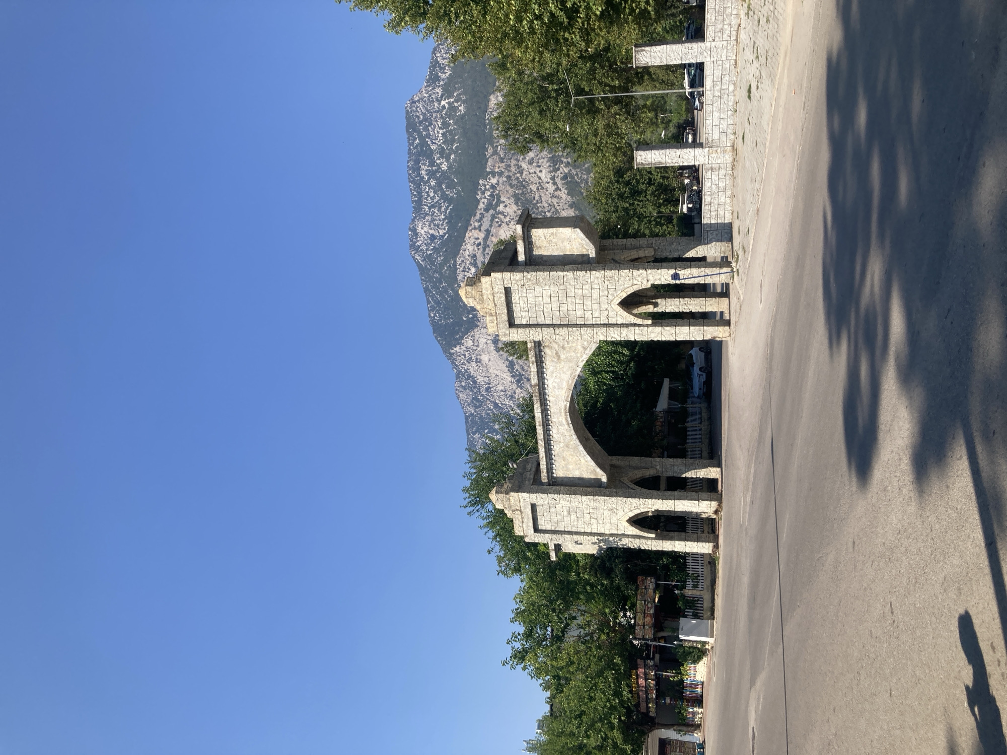 An archway made of light-colored stone stands in the foreground, featuring intricate carvings and two smaller arched openings on either side. Beyond the arch, a tree-lined street is visible with several buildings and parked cars. In the background, a majestic mountain range dominates the landscape under a clear blue sky. This scene is located in Kemer, Turkey, a popular tourist destination known for its natural beauty and historical sites.