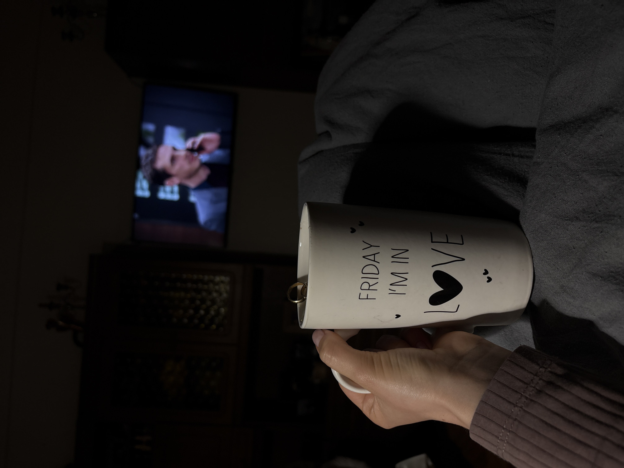 A dimly lit indoor scene shows a person holding a white mug with the text "Friday I'm in Love" and a heart symbol, containing a gold ring inside. A television screen in the background displays a man's face in a suit. A dark, patterned cabinet is visible beneath the TV. The person is wearing a long-sleeved shirt. The environment appears relaxed, suggesting a casual setting indoors. The scene lacks any evidence to connect it to the city of Minsk, Belarus.