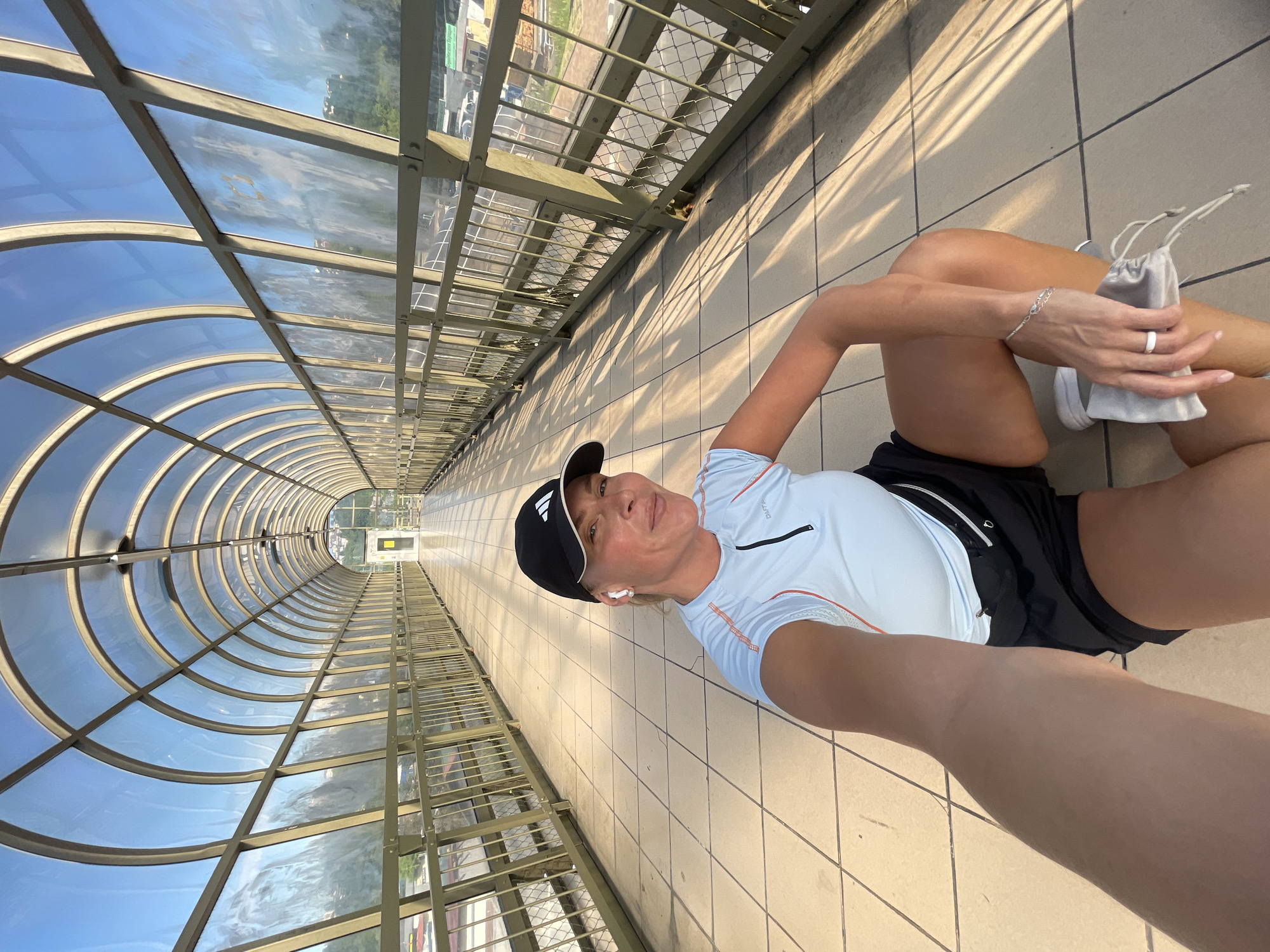 A woman is captured in a selfie, lying or resting on the tiled floor of a modern, glass-enclosed pedestrian walkway in Sochi, Russia. The photo is oriented with her head to the left and feet to the right, looking up at the camera she holds. She appears relaxed, with a slight smile on her face.
She is dressed in athletic wear, including a black baseball cap featuring a white Adidas logo, a light blue short-sleeved athletic shirt with "CRAFT PRO" visible and orange trim, and black athletic shorts. A white wireless earbud is visible in her left ear. Her left wrist displays a silver chain bracelet, and a white ring is on her left ring finger. In her left hand, she holds a small, light grey drawstring pouch.
The walkway is a striking architectural feature, characterized by a long, arched, tunnel-like structure. Its ceiling and upper sides are composed of clear and translucent glass panels supported by metal frames, through which a bright blue sky is visible. Sunlight streams through, casting prominent, geometric shadows of the structure onto the light-colored tiled floor, suggesting a clear and sunny day, likely late morning or afternoon. Through the side panels, hints of an urban or suburban environment can be seen. The long perspective of the tunnel recedes into the distance, emphasizing its length.