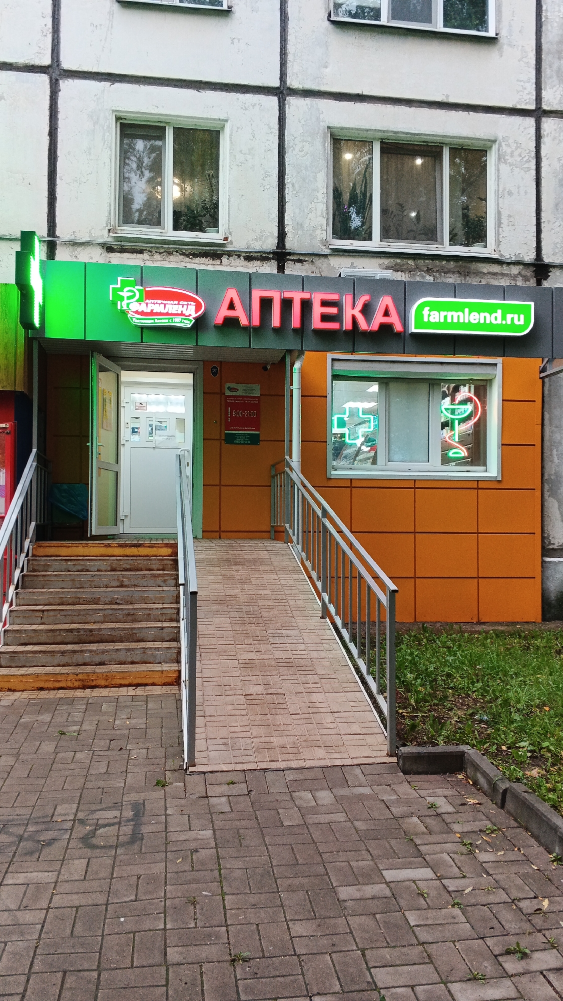 The image shows the entrance to an "APTEKA" (pharmacy) in Izhevsk, Russia. The pharmacy has a prominent green and red sign with "APTEKA" written in large red letters and "farmlend.ru" in green. A green cross symbol is also visible on the left side of the facade and illuminated in the window on the right. The entrance features a set of stairs to the left and a ramp to the right, both leading up to the doorway. The building appears to be a multi-story apartment complex with white-gray facade. The ground is paved with stone tiles. A sign on the wall inside the entrance indicates the operating hours are from 8:00 to 21:00. The scene is an exterior shot of a commercial establishment in a residential area.