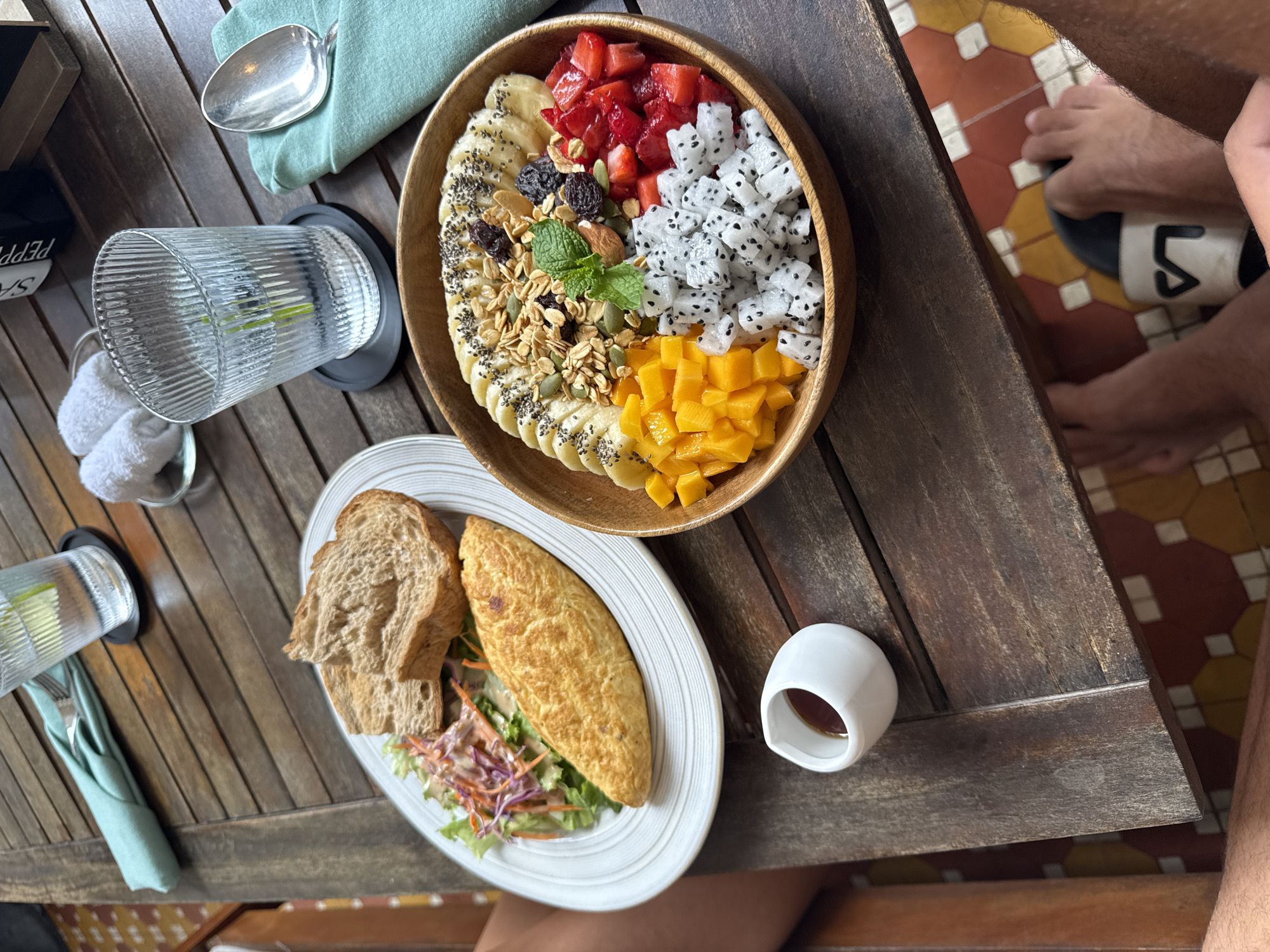 A high-angle shot shows a wooden table with two plates of food, drinks, and utensils. A wooden bowl contains a fruit arrangement including bananas, strawberries, dragon fruit, mango, granola, and raisins. The plate below contains an omelet, salad, and two slices of bread. A small white cup is next to the plate. A water glass with lime is on the table with a silver spoon on a light green napkin. An adjacent floor shows tiled flooring in red, white, and yellow. Part of a person wearing a black-and-white shoe is visible on the right. The image is consistent with a breakfast setting. The location in Đà Nẵng, Vietnam, is inferred from the style of the food and overall ambiance.