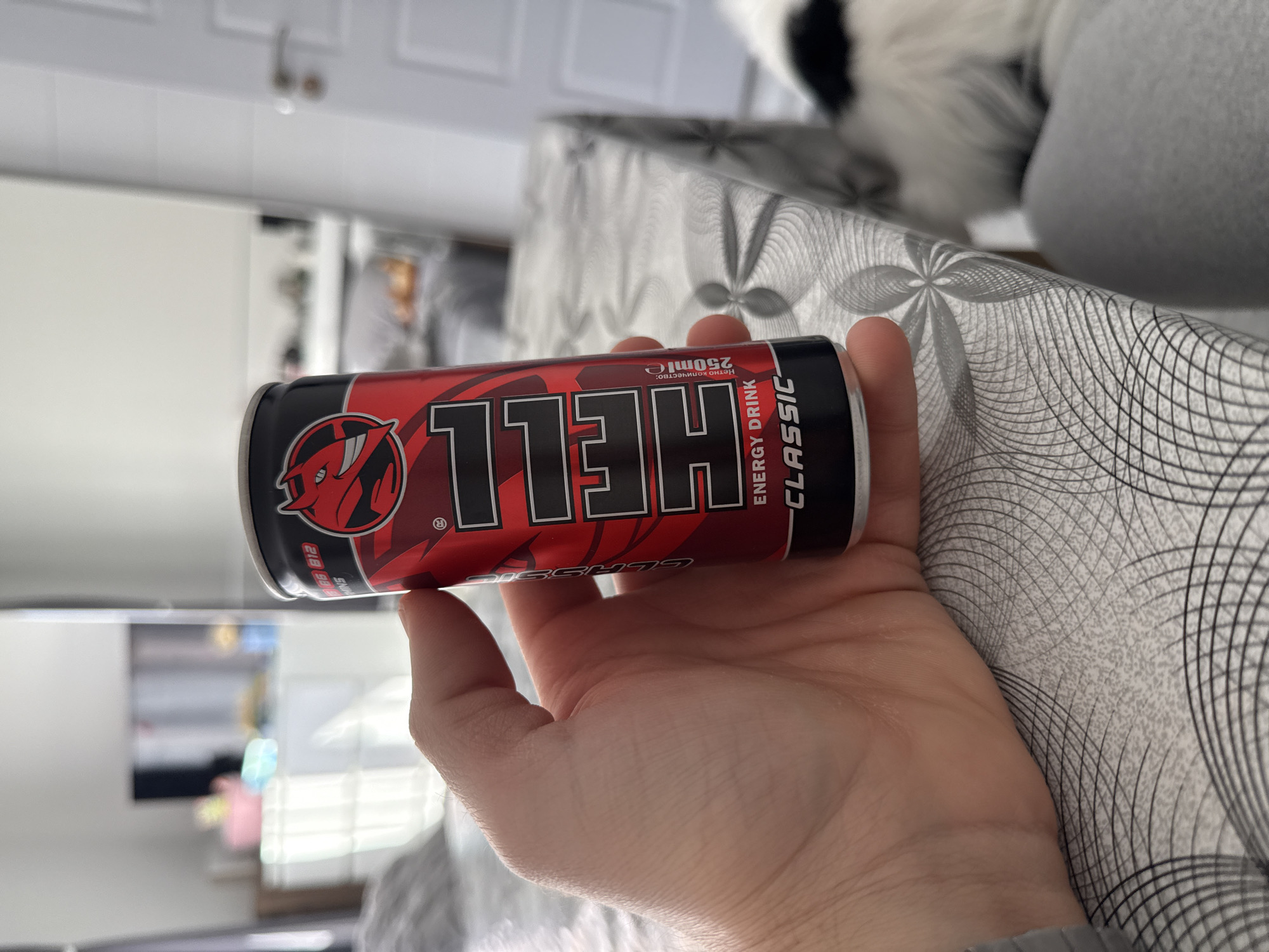 A person is holding a red and black can of "HELL" energy drink, "Classic" flavor, in their right hand. The can displays a red devil logo. The background includes a light-colored surface with a gray geometric pattern. In the blurry background are what appears to be interior rooms, including a white door with silver hardware and a television. The location is not discernible as Stara Zagora, Bulgaria based on the image content alone.