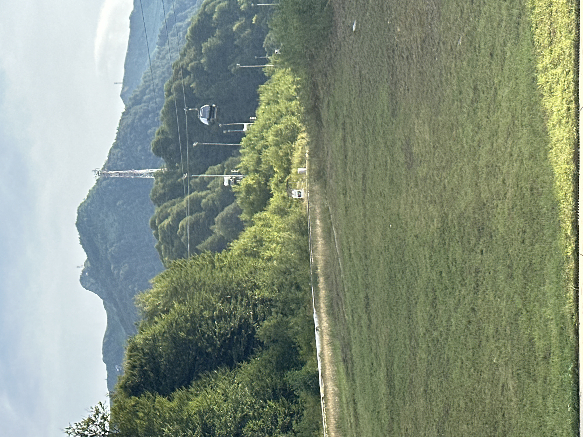 The image depicts a mountainous landscape with lush green vegetation and a clear sky. In the foreground, a grassy slope dominates the lower half of the frame, transitioning to a path or road with a railing at the mid-ground. Beyond this, a dense forest covers the slopes of rolling hills. In the distance, a prominent transmission tower with red and white accents stands atop a mountain, and another smaller tower is visible further to the right. A cable car, a single gondola, is suspended from wires in the mid-ground, traversing the scene. The environment suggests a recreational area, possibly a ski resort or a mountain park, given the presence of the cable car and the manicured grassy slope. The overall scene is peaceful and natural, with man-made structures integrated into the landscape.
