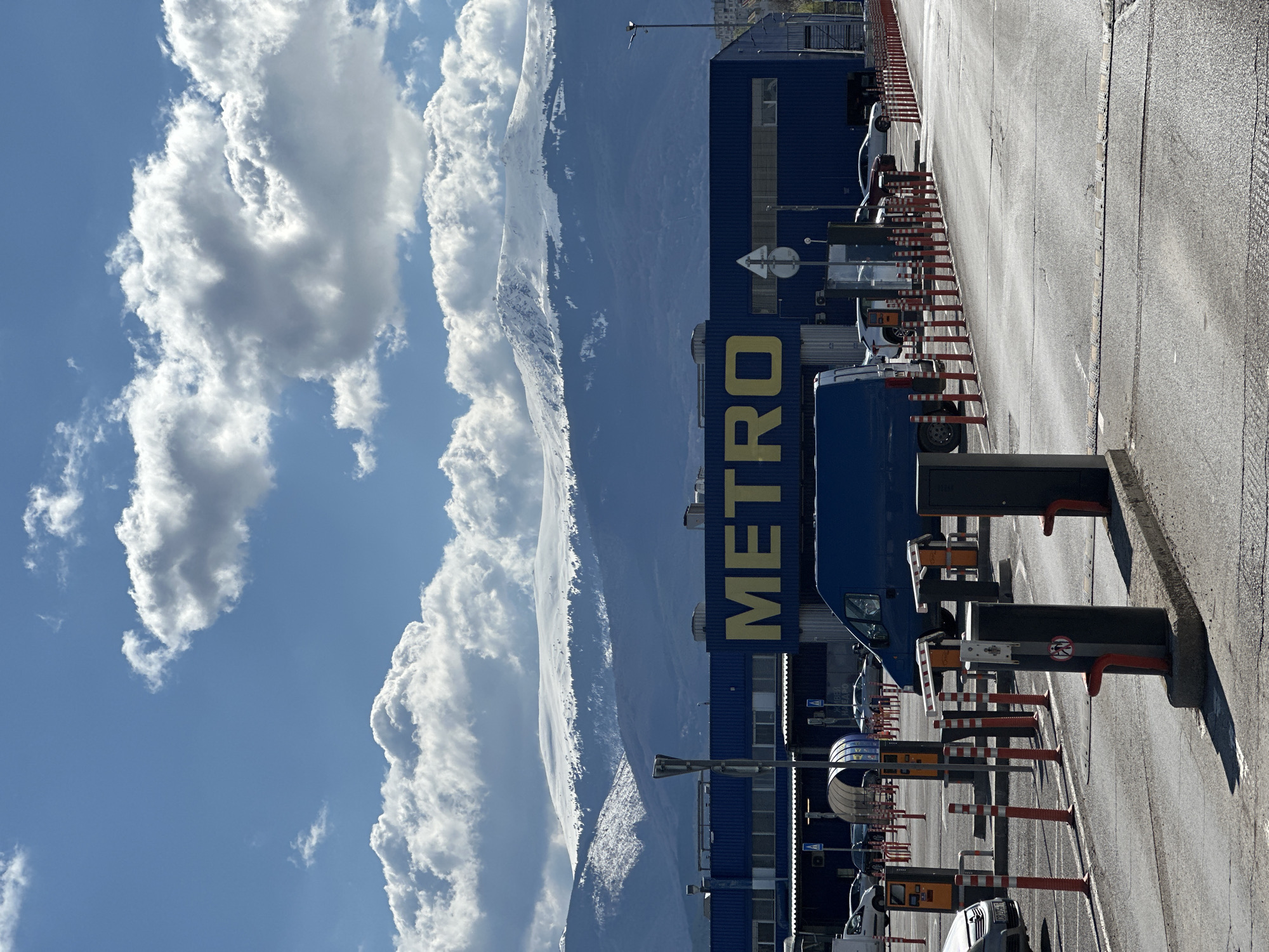 The image shows the METRO Cash & Carry store in Sofia, Bulgaria, with mountains in the background partially obscured by clouds. The blue building with the large yellow "METRO" lettering is visible from its parking lot entrance. The parking lot is marked by red and white barriers and automated entry/exit gates. Several vehicles are parked within the lot. The day is sunny with some cumulus clouds in the sky. The image is taken at ground level, looking up at the building and mountains.