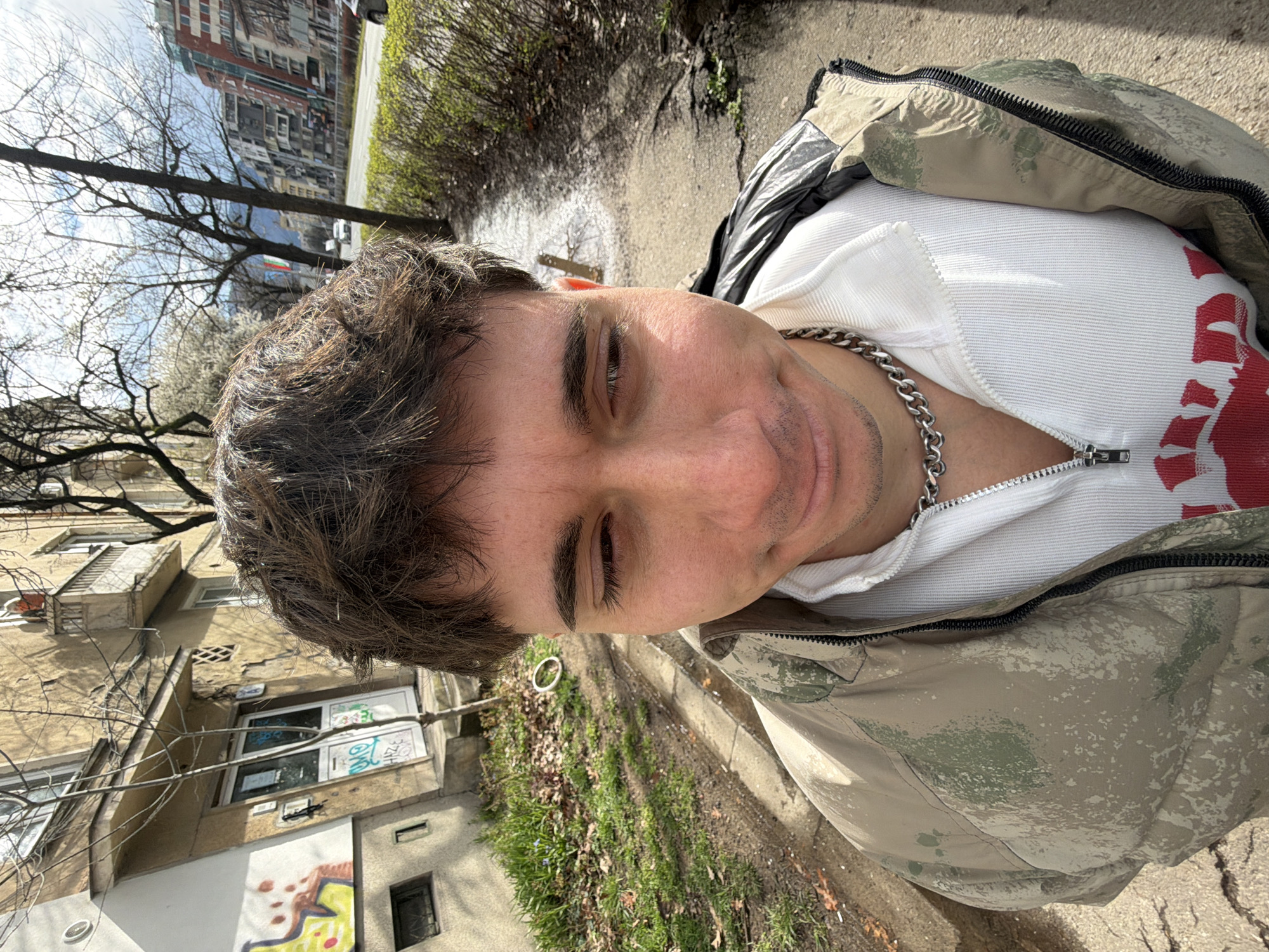 A man is pictured outdoors, looking down at the camera, wearing a white shirt partially zipped open with a silver chain necklace, and a camouflage jacket. He appears to be on a sidewalk with a building to his left and vegetation alongside the path. Other buildings are visible in the background with bare trees. The photograph was taken during the daytime. The specific location is likely in Sofia, Bulgaria, judging by the architectural style of the buildings in the background, with their distinctive concrete and plaster facade.
