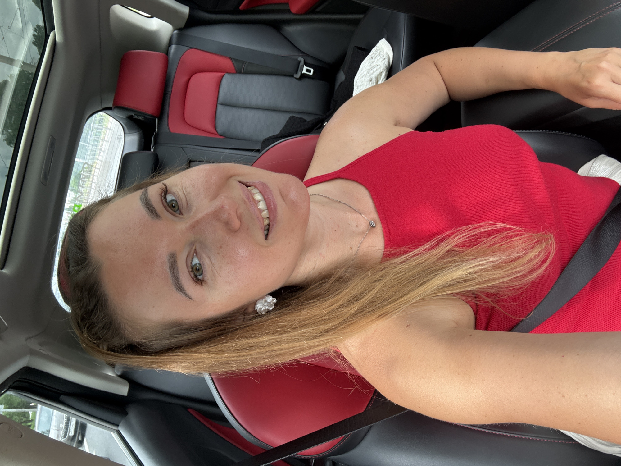 A medium shot captures a young woman with long blonde hair, wearing a red tank top and a silver necklace, as she takes a selfie inside a car. The car's interior features black leather seats with red accents and a sunroof. The woman is smiling, with her eyes looking towards the camera. She is seated in the passenger seat, and the driver's side of the car, with a black seat belt visible, is in the background. Outside the car, a gray vehicle is seen on a road, with some greenery and fencing in the distance, suggesting an urban or suburban environment. The lighting is natural, indicating it is daytime.
