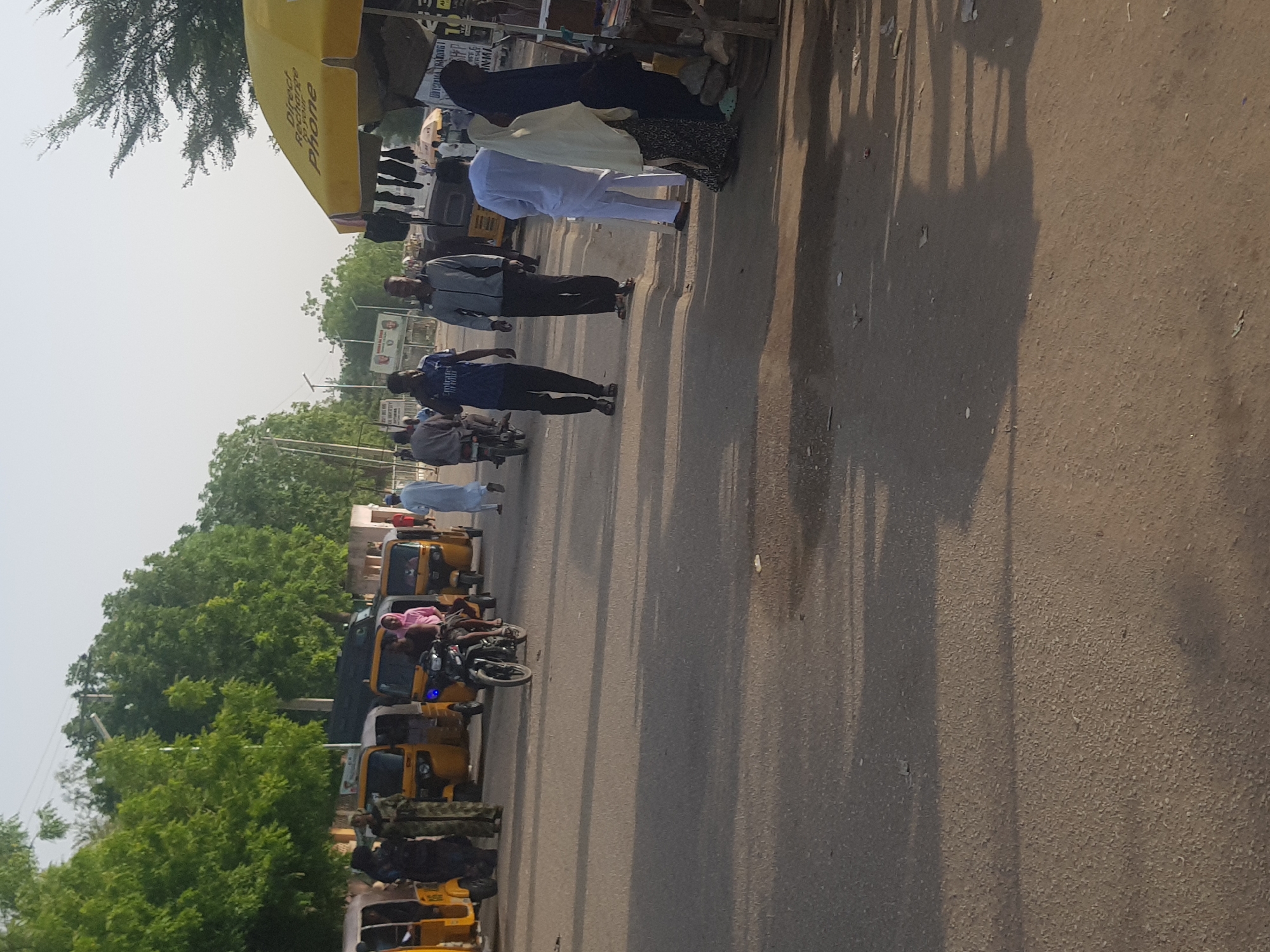 The image shows a street scene in Dabawa, Nigeria, with several people walking along the road and a row of yellow three-wheeled vehicles parked along the side. Several men are walking in different directions. Some people are standing near a yellow canopy, possibly a vendor stall. Trees line the opposite side of the road from the parked vehicles. The sky is light, indicating it is daytime. The road surface is paved and there are shadows cast on the road.