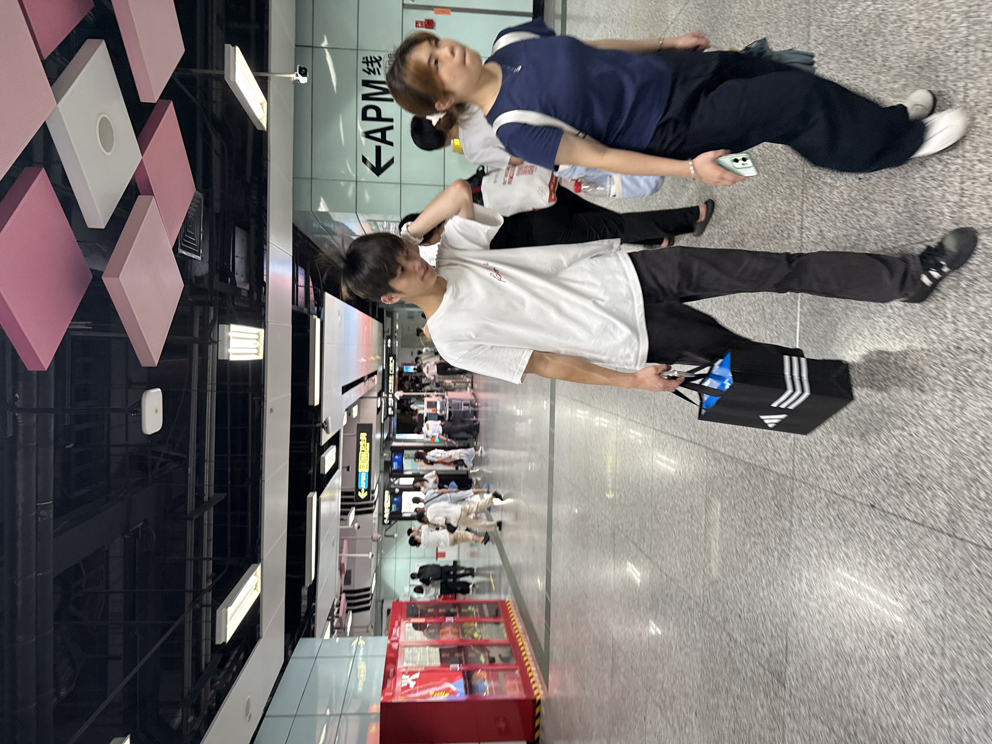 The image depicts a group of people in a modern, well-lit indoor environment, likely a subway station or a similar public transit hub. The scene is dominated by a polished, speckled gray floor and sleek, contemporary architecture. The subjects are a mix of genders and ages, with several individuals walking and standing.
In the foreground, a young man wearing a white t-shirt and black pants is walking, carrying a black Adidas bag. To his left and slightly behind him, a woman in a dark blue t-shirt and black pants is also walking, holding a smartphone. Behind them, other people are visible, some walking in the same direction, others standing or waiting. The background reveals more of the station's infrastructure, including directional signage, display screens, and what appears to be a vending machine or kiosk.
Notable details include the clean and modern aesthetic of the location, suggesting it is a relatively new or well-maintained facility. The lighting is bright and even, indicating it is daytime, although there is no direct view of the outside weather. The general atmosphere seems to be one of movement and everyday transit.
Visible text includes "APM线" in Chinese characters and English letters on a green sign pointing left, indicating the direction of the APM line. There is also some Chinese text on other signs further in the background. The overall impression is a busy, functional public space.