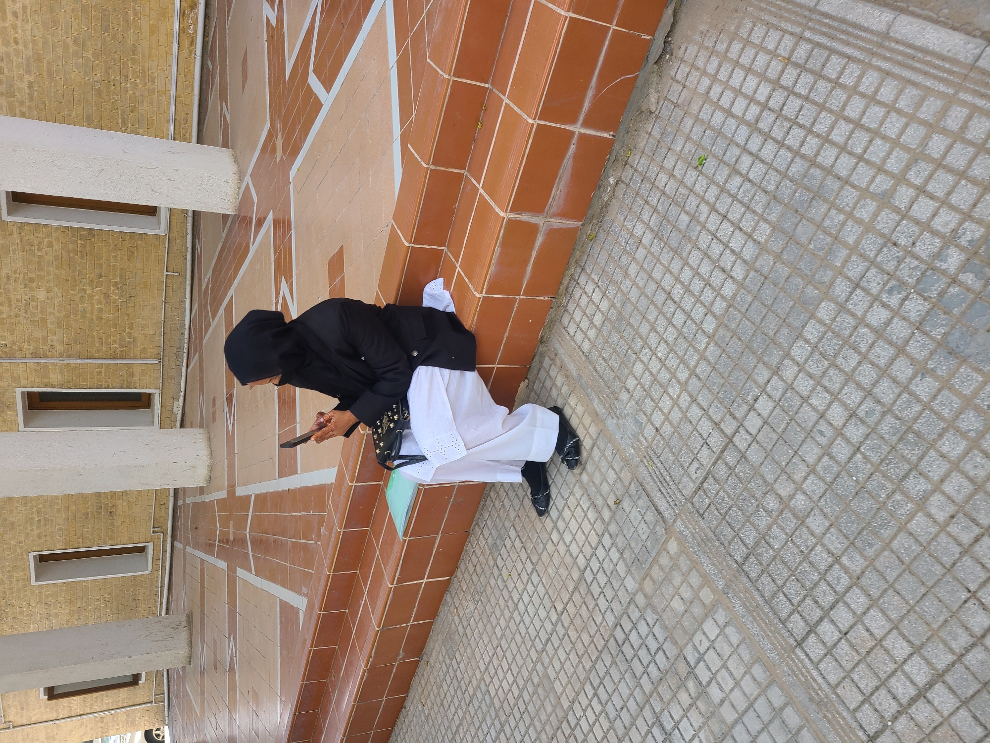 A woman in a black hijab and jacket and white trousers is squatting down, using a smartphone. She is in Quetta City Tehsil, Pakistan. She sits on a gray tiled sidewalk with a red tiled wall beside it.