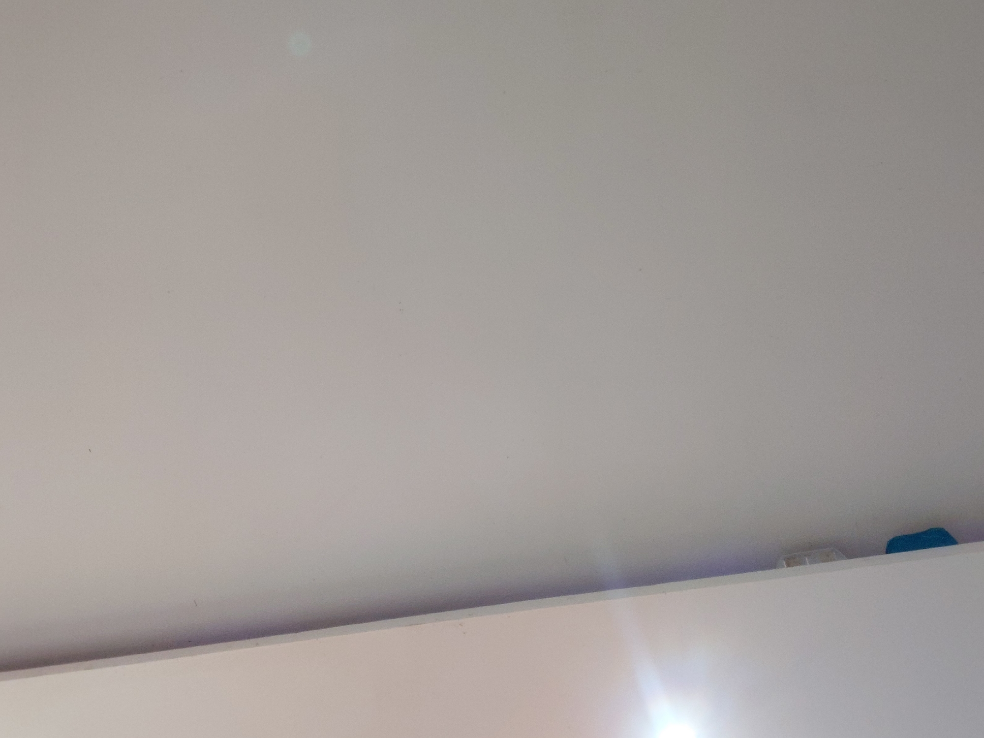 The image displays an interior architectural detail, likely a ceiling or the upper portion of a wall, in Santa Ana, Venezuela. The dominant feature is a smooth, light-colored surface, appearing off-white or very light grey, which occupies most of the frame. A faint, greenish, circular reflection is visible in the upper-left quadrant of this surface.
A horizontal line, possibly a corner where a wall meets a ceiling or another wall, runs across the lower part of the image, separating the main light surface from a slightly darker, lighter-colored plane at the very bottom.
On the right side of the image, peeking just above this lower plane, are two small objects. One is a light-colored, rectangular item, possibly a container or part of a fixture. To its immediate right, a small, irregularly shaped blue object is visible. A strong lens flare originating from a bright light source is present in the bottom right corner of the frame, indicating a well-lit environment, possibly due to natural light from a window or a powerful artificial light. No people are visible, and no text is present in the image.