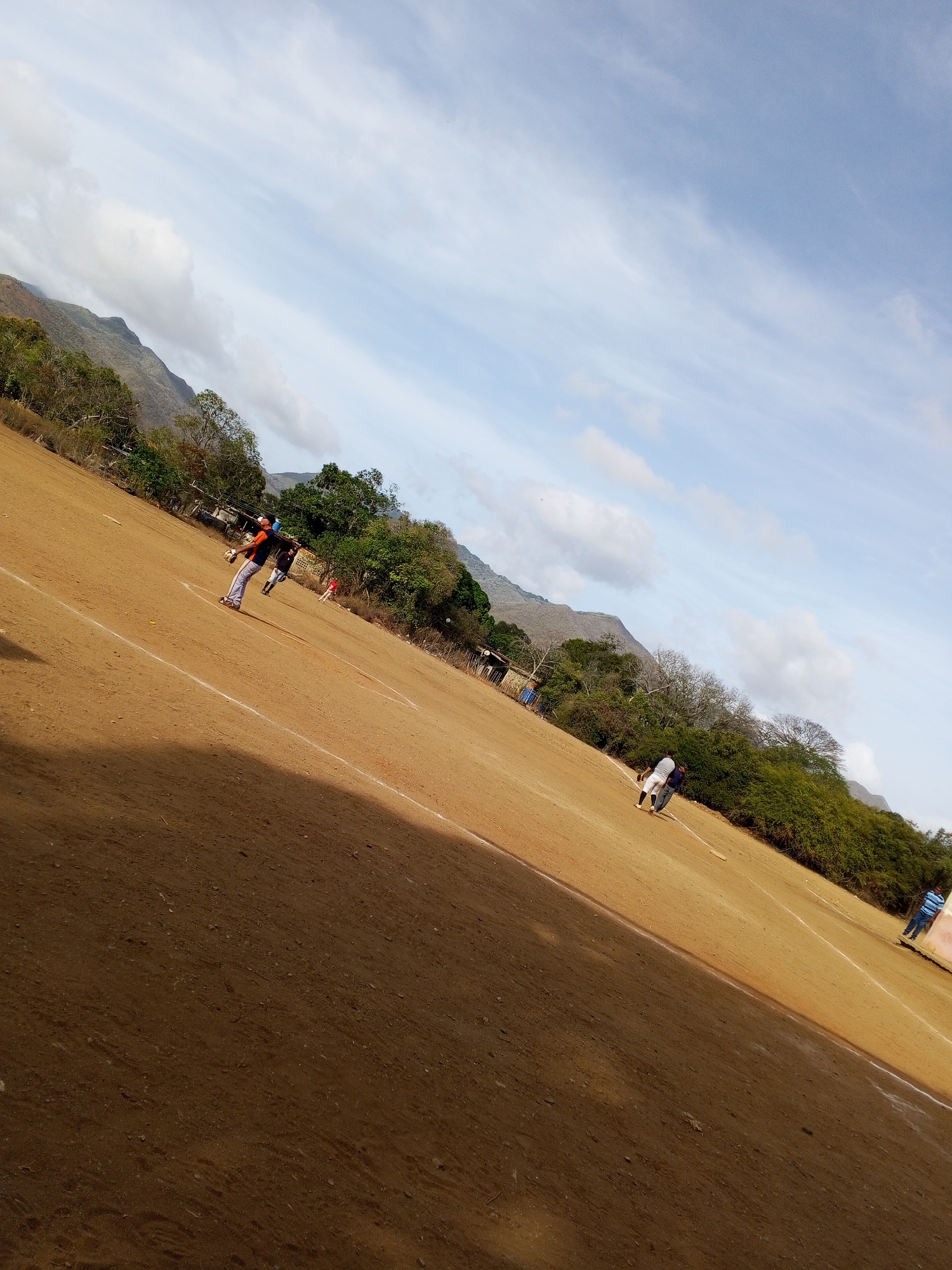 A baseball field located in Unknown, Unknown, with a dirt infield and a green outfield, is the setting for the image. The perspective is angled, showing a large portion of the dirt infield on the left. Mountains rise in the background under a partly cloudy sky. Several people are scattered on the field. One individual, located on the first baseline, is wearing an orange and black jersey and gray pants. Other individuals, either players or spectators, are further out in the outfield. The lighting is bright, casting shadows on the field.
