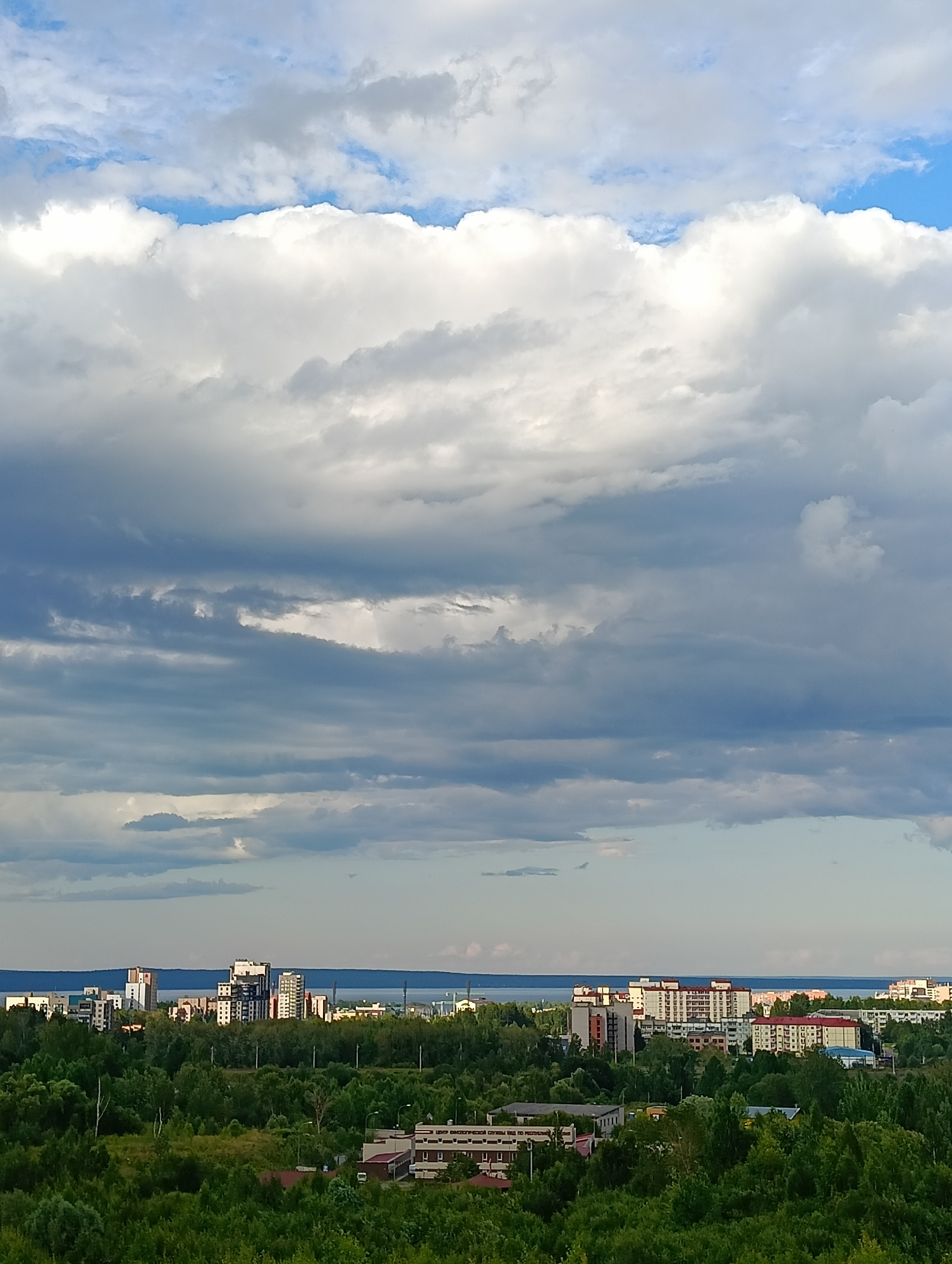 The image captures a city skyline with a prominent body of water in the background, likely Lake Onega, a significant feature of Petrozavodsk, Russia. A dense expanse of green trees forms the foreground, with various buildings of different heights and architectural styles interspersed within and beyond the treeline. These buildings appear to be residential and possibly commercial structures. The sky is filled with a dramatic arrangement of clouds, ranging from bright white cumulus formations to darker, more layered stratus clouds, indicating a dynamic weather pattern. The overall scene suggests an urban environment integrated with natural elements, under a vast, cloudy sky.